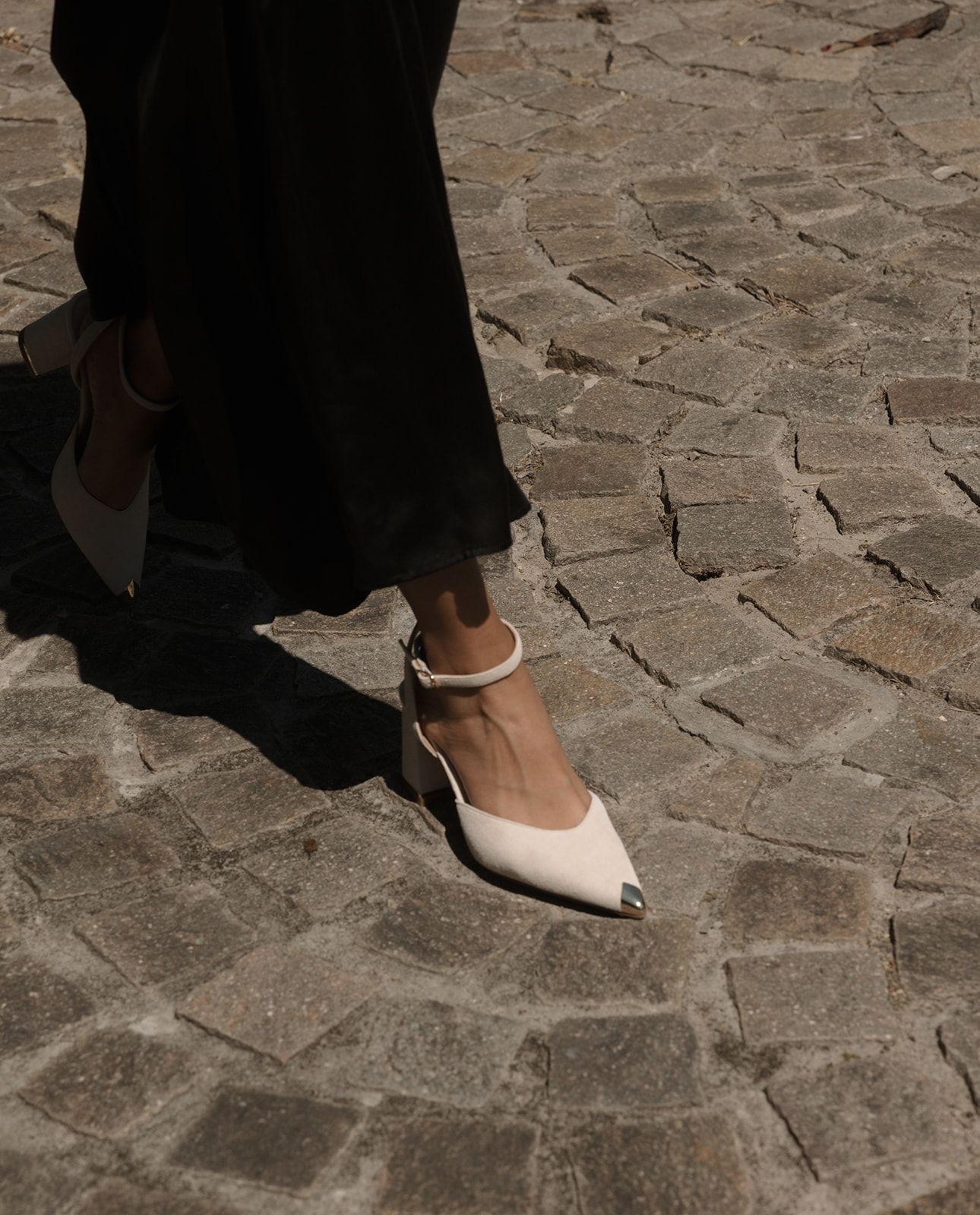 A person wearing black clothing and white ballet flats walks across a cobblestone street.