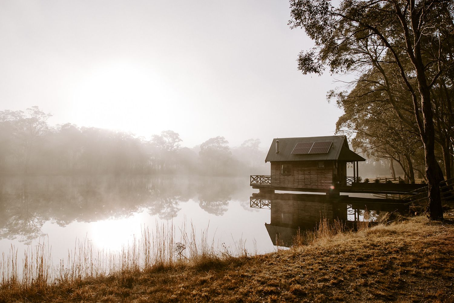 Misty lakeside boathouse on a calm morning with fog rolling over still water, surrounded by autumn trees and golden grass.