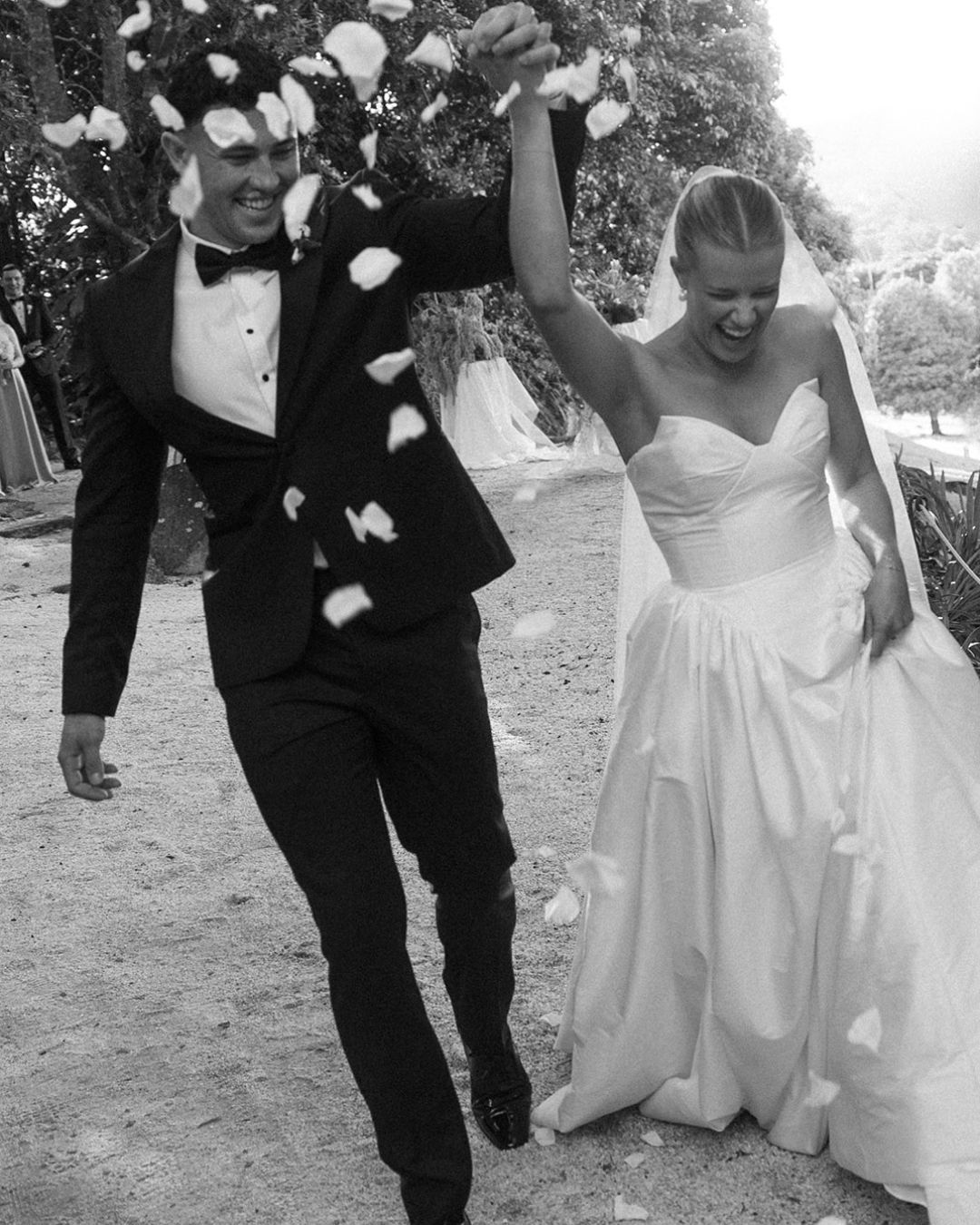 Black and white photo of a joyful bride and groom walking together as flower petals rain down on their wedding day.