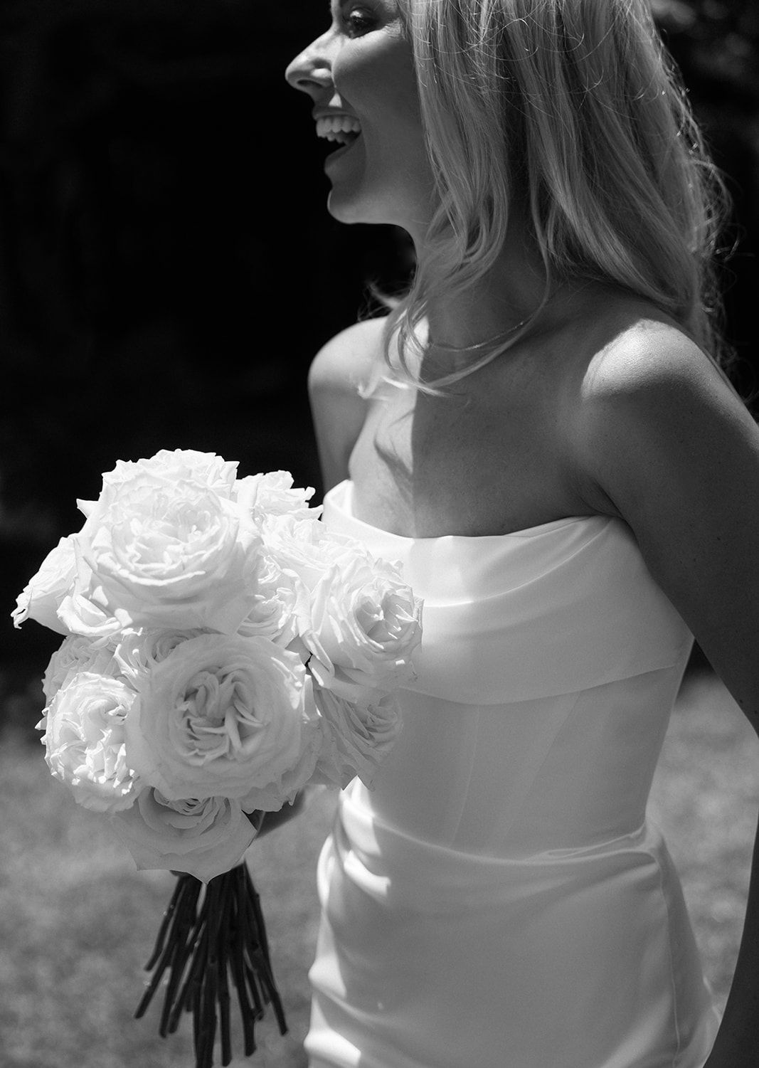 A bride in a strapless white gown holds a bouquet of white roses in black and white photography.