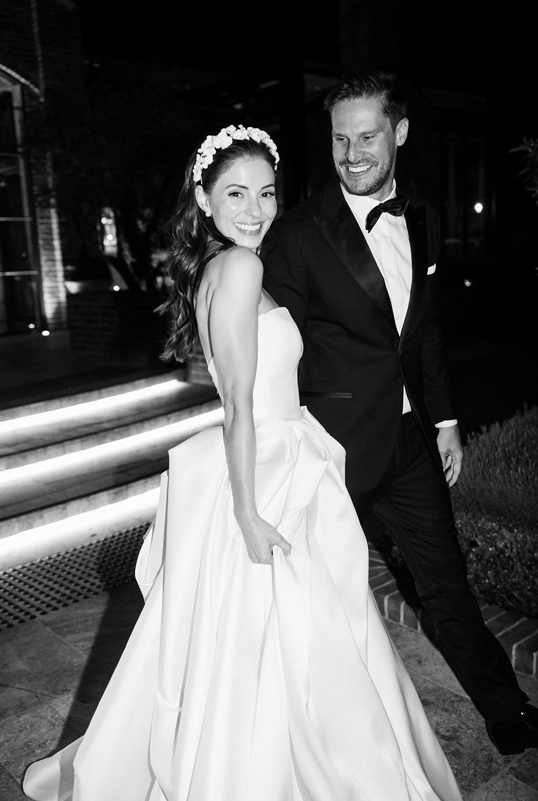 Black and white wedding photo showing a bride in an elegant white gown with a floral crown and groom in classic tuxedo.