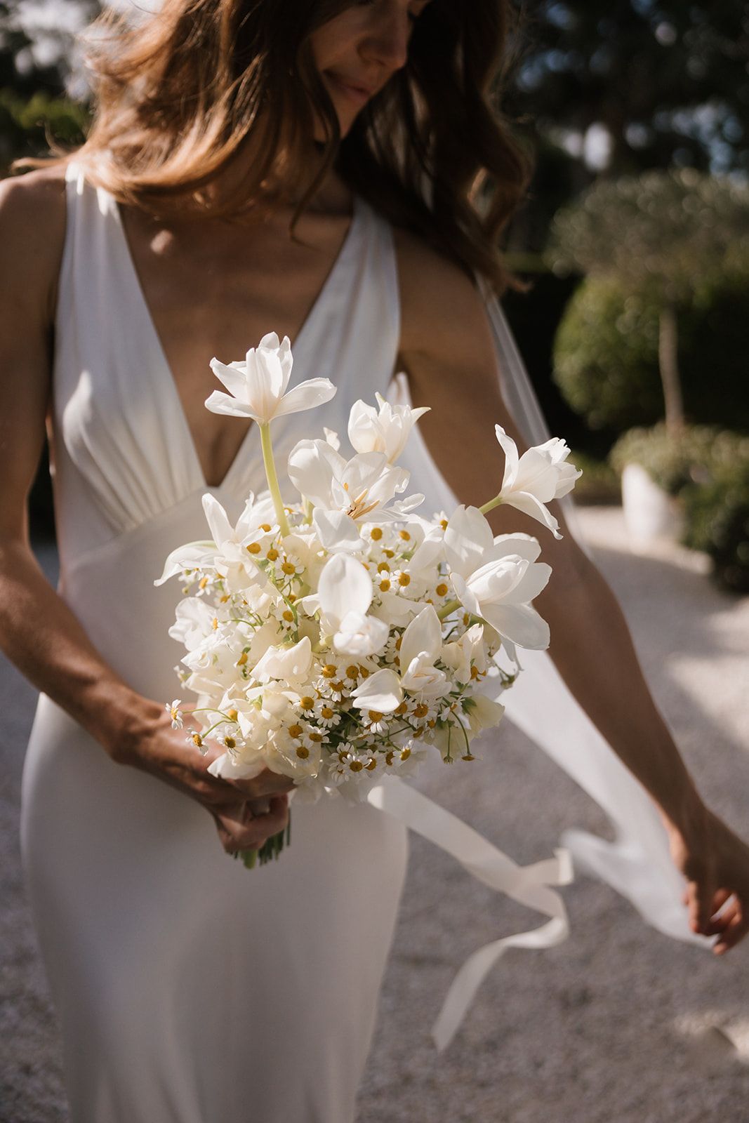 Three bridesmaids in elegant white dresses hold beautiful bouquets of white flowers and greenery in outdoor setting.