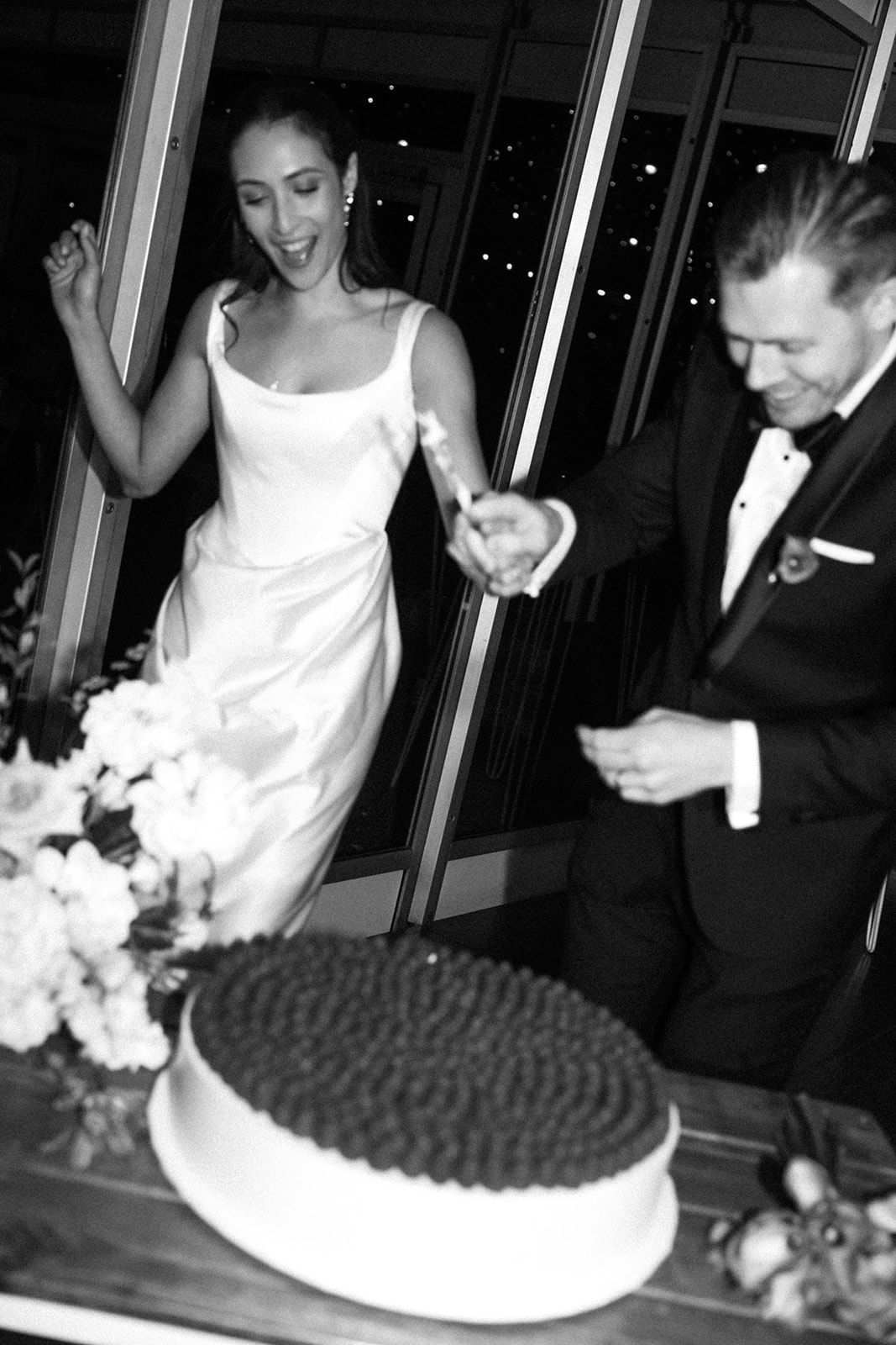 Black and white photo of a bride in a white dress reacting with delight as a groom cuts into their wedding cake.