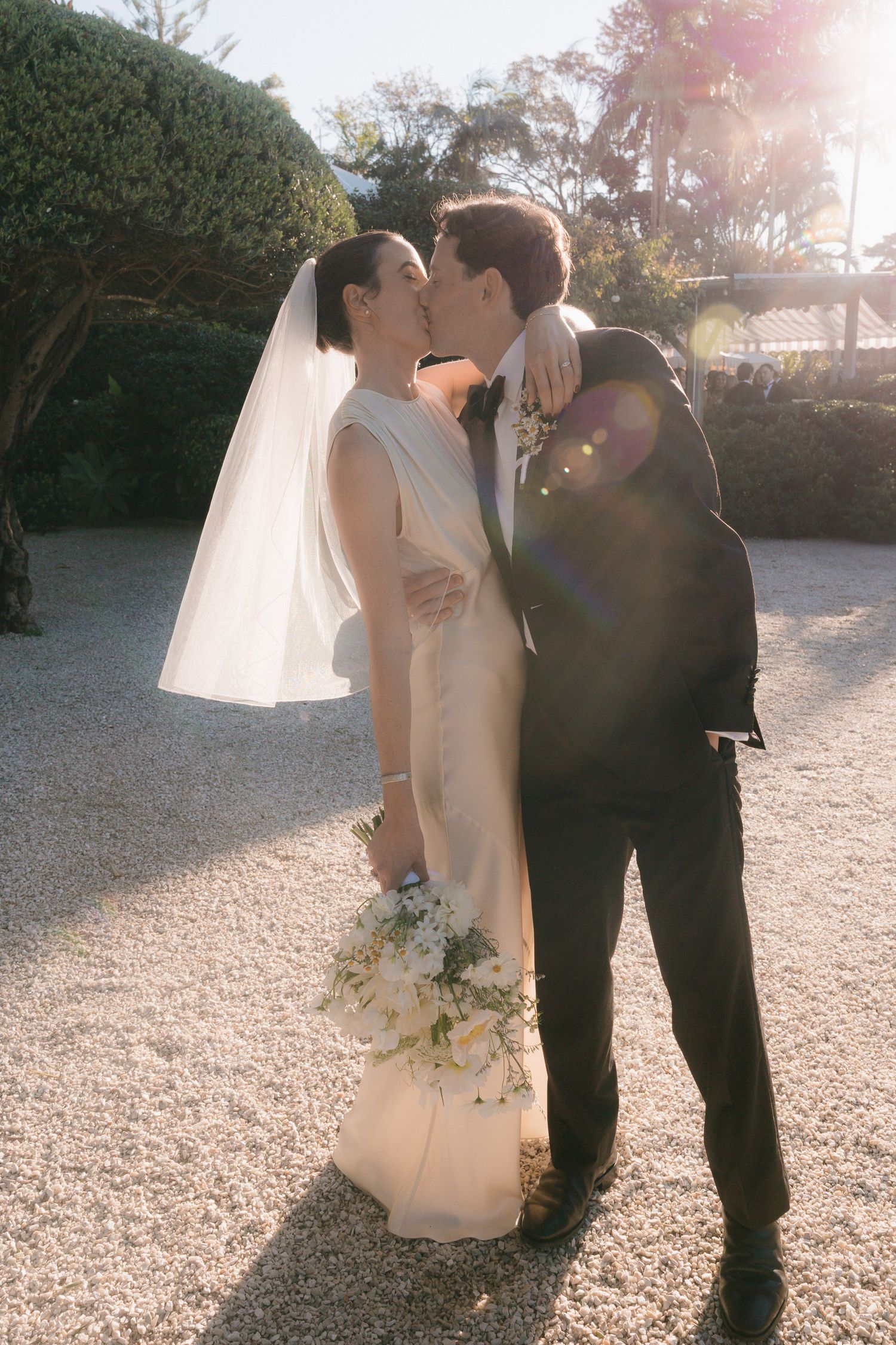Sun flare illuminates a romantic wedding couple embracing outdoors on a gravel path during their photoshoot.