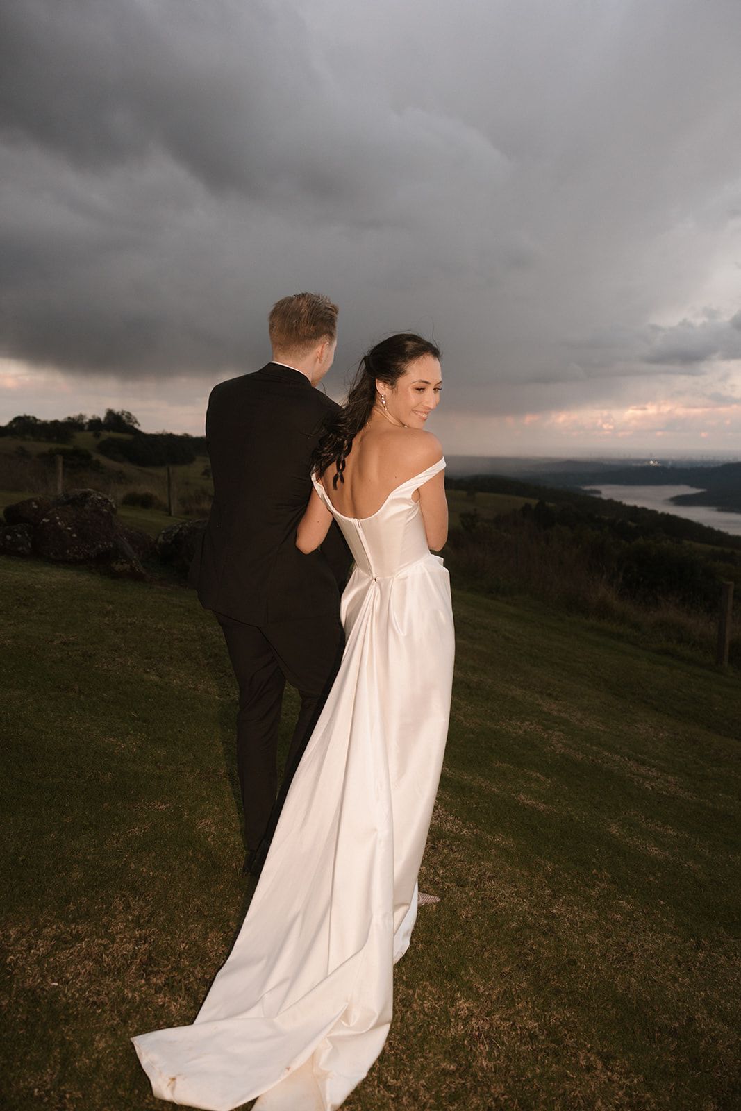 A couple in wedding attire stand on a hilltop during a dramatic sunset with storm clouds overhead.
