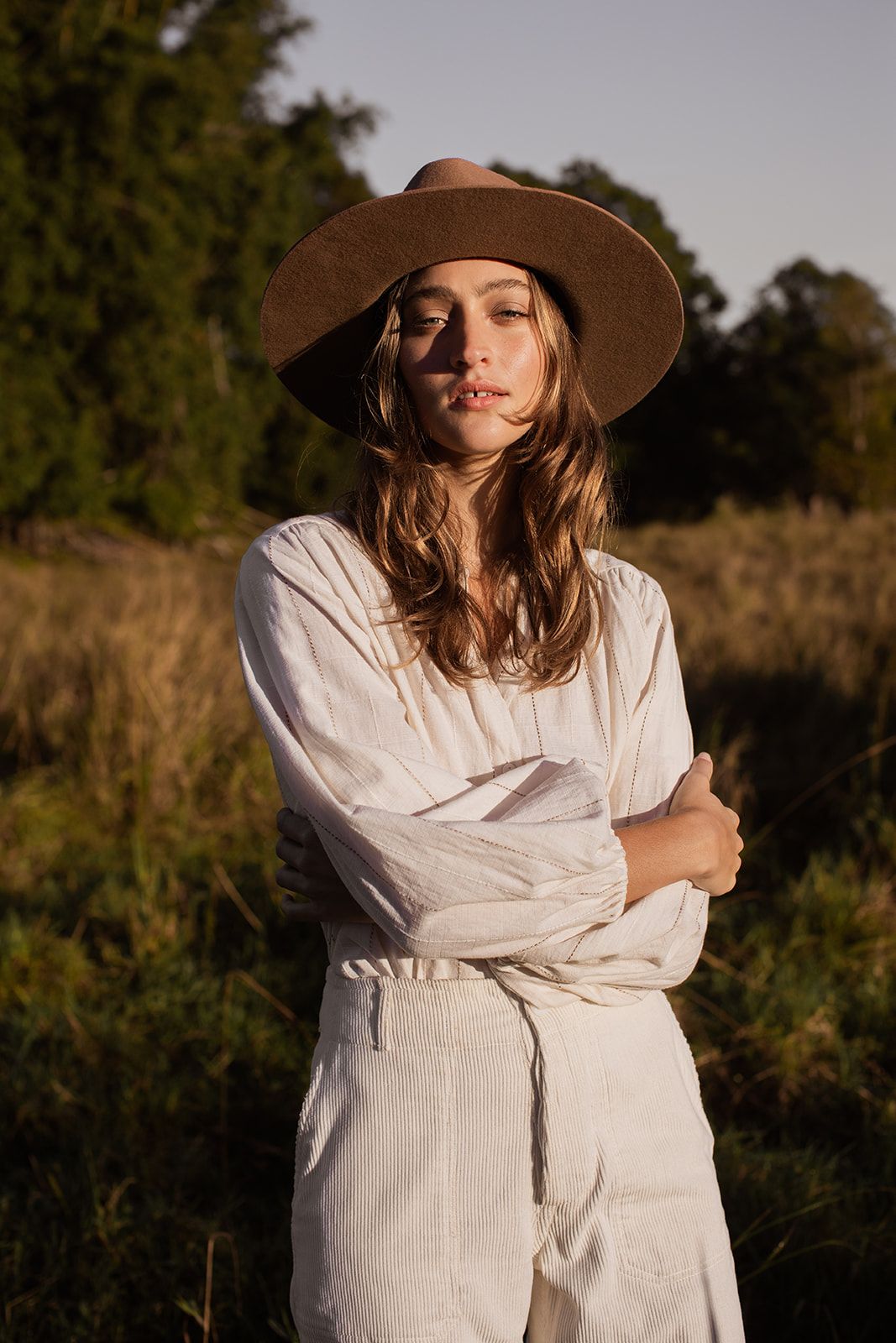 Woman in white linen dress and wide-brim hat standing in golden hour sunlight in a natural field setting.