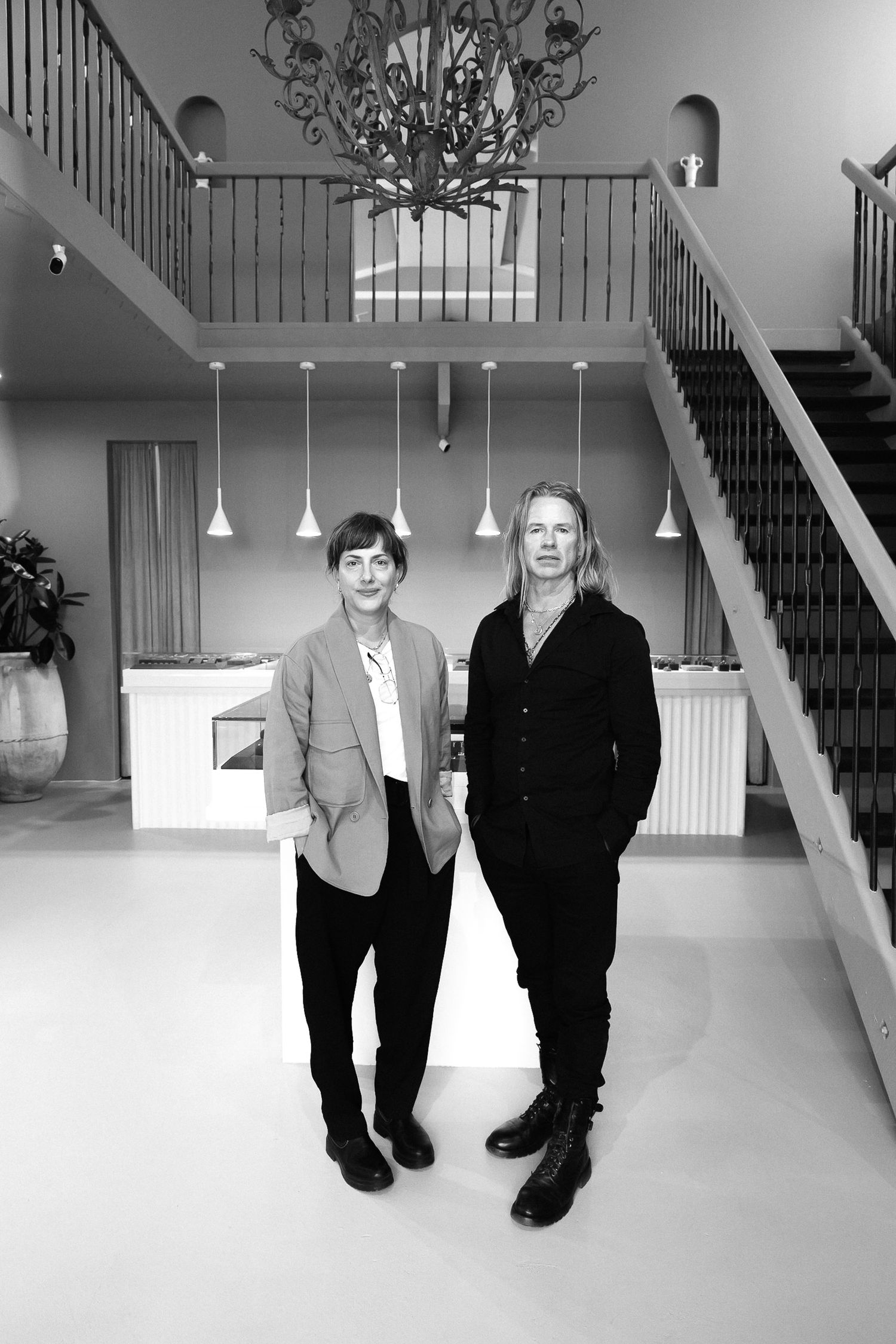 Black and white interior photo showing two people standing in an elegant foyer with pendant lights and a staircase.
