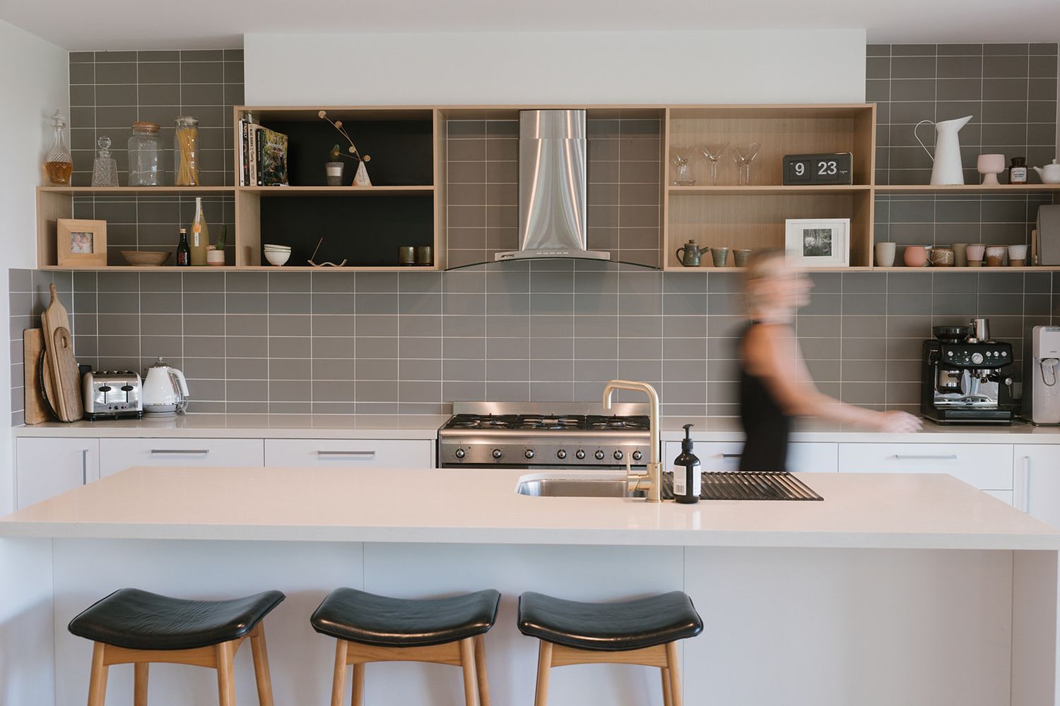 Modern kitchen interior with wooden cabinets, gray tile backsplash, and black bar stools at white countertop peninsula.