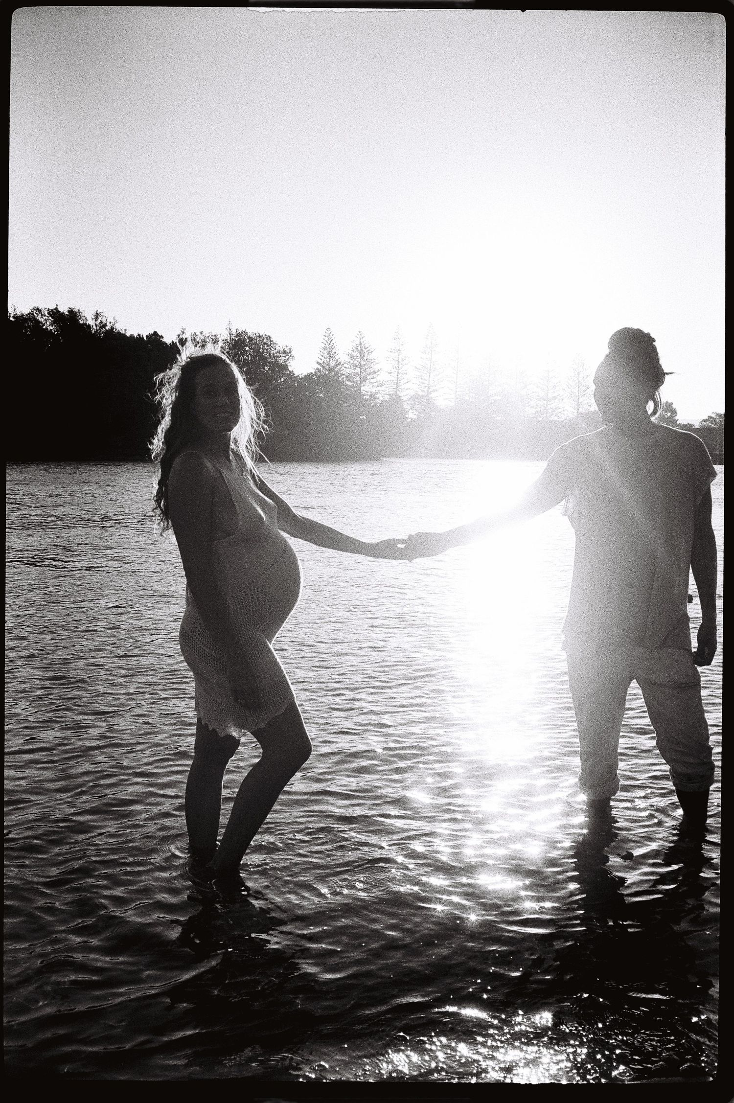 Black and white photo of a pregnant woman and partner holding hands in a sunlit lake, trees in background.