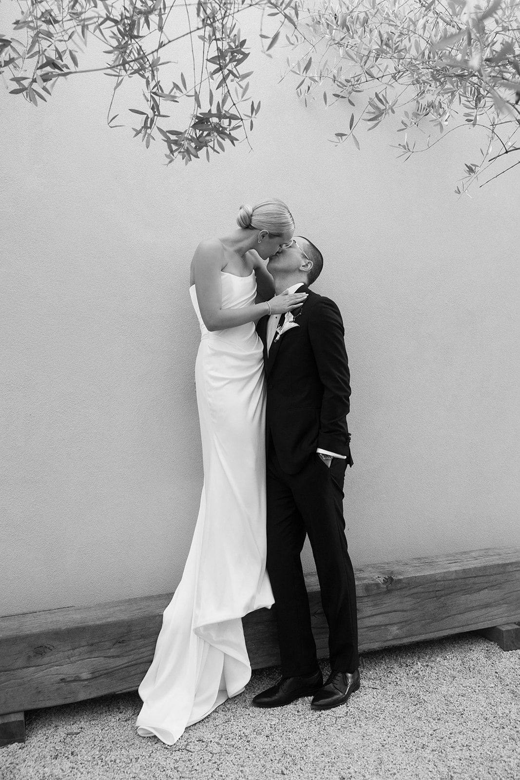 Black and white photo of a bride and groom sharing a kiss in wedding attire against a neutral wall.