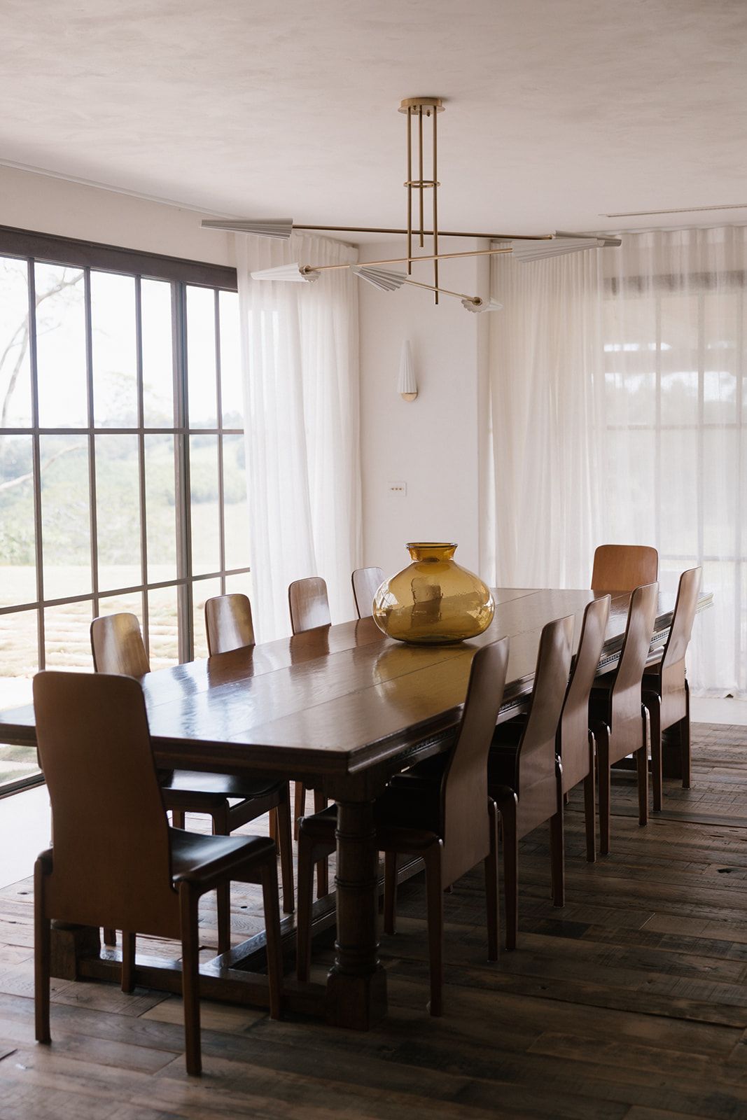 Modern dining room with long wooden table, upholstered chairs, brass light fixture and amber glass vase centerpiece.