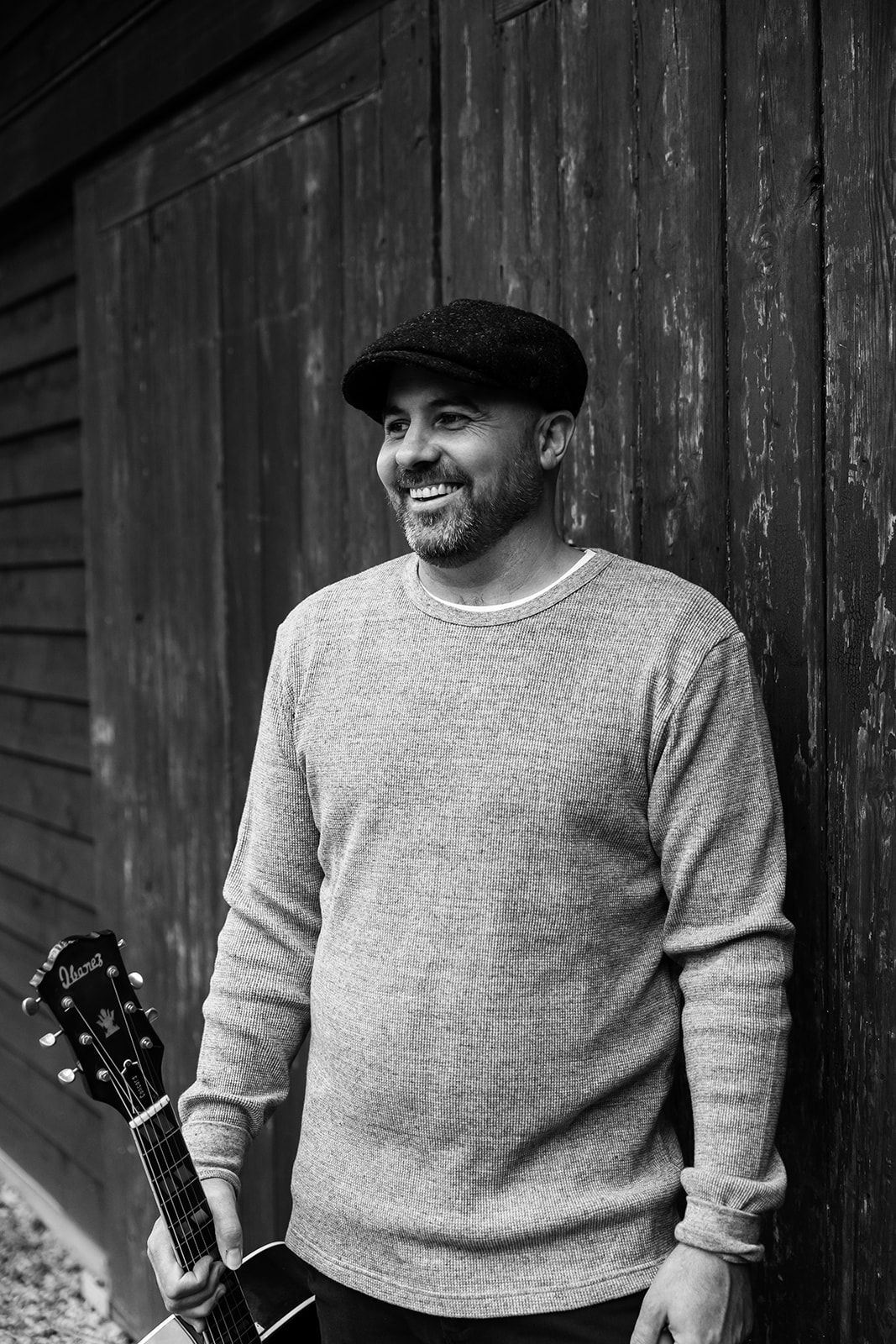 A person wearing a gray sweater stands against a dark wooden wall while holding an acoustic guitar.