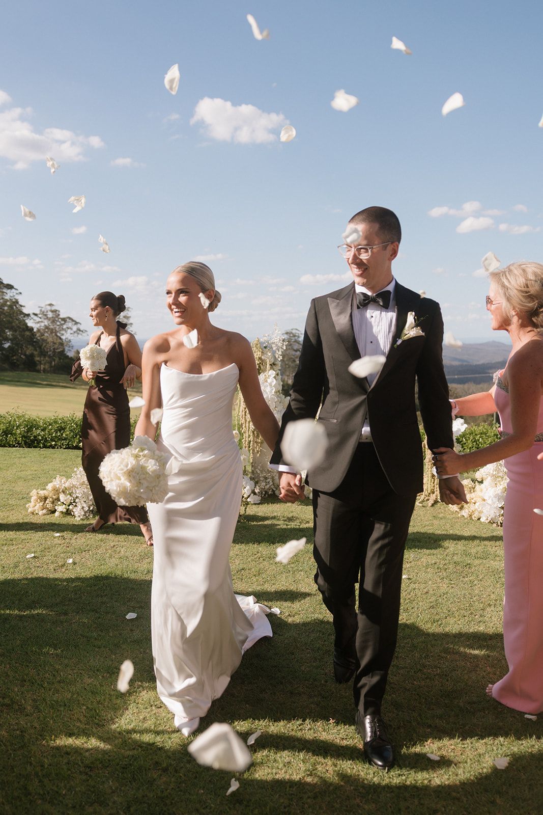 Bride and groom walk through flower petal confetti after outdoor wedding ceremony on lush green lawn.