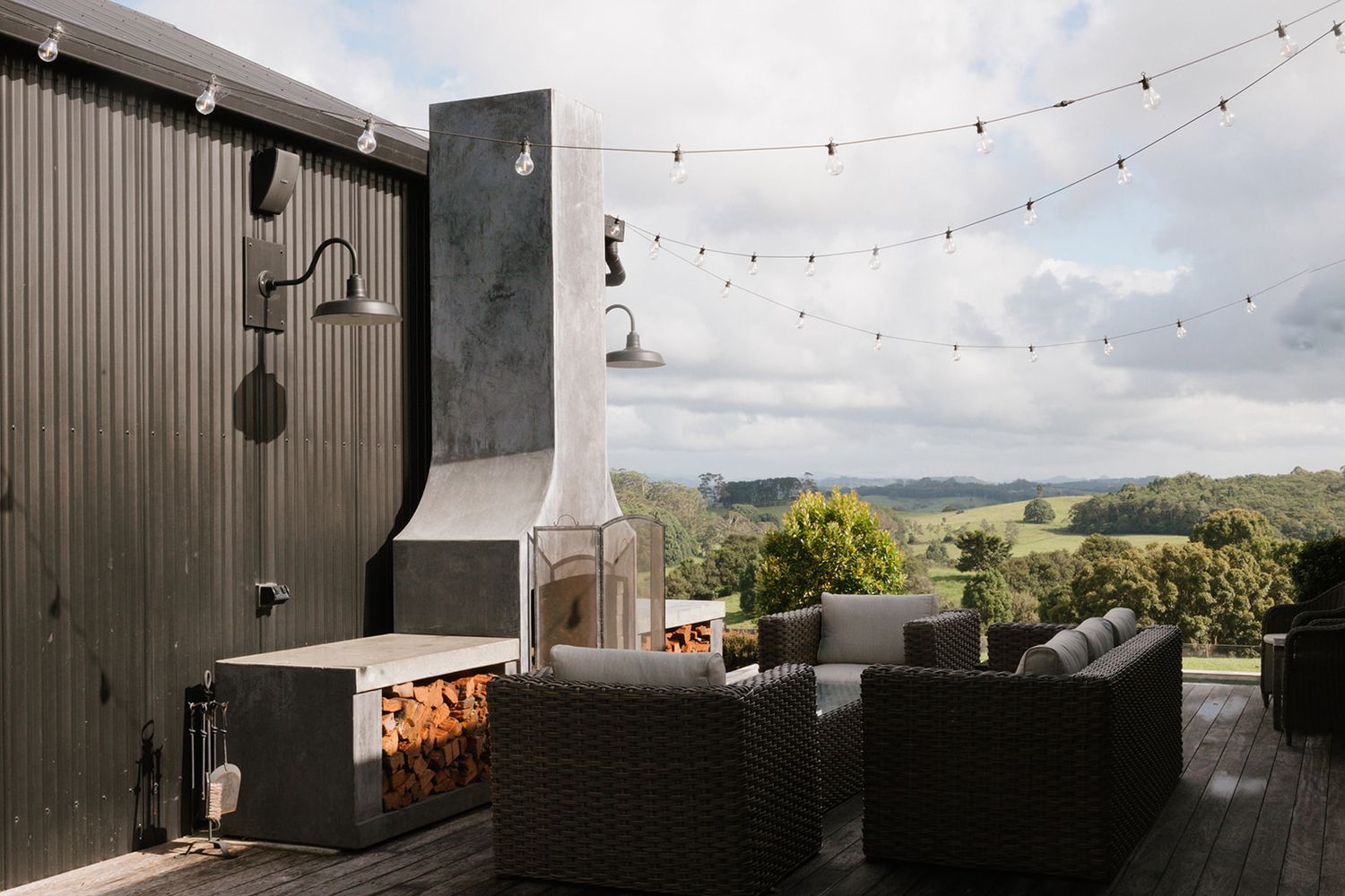 Outdoor deck with wicker furniture, concrete fireplace, string lights, and scenic green hills view.