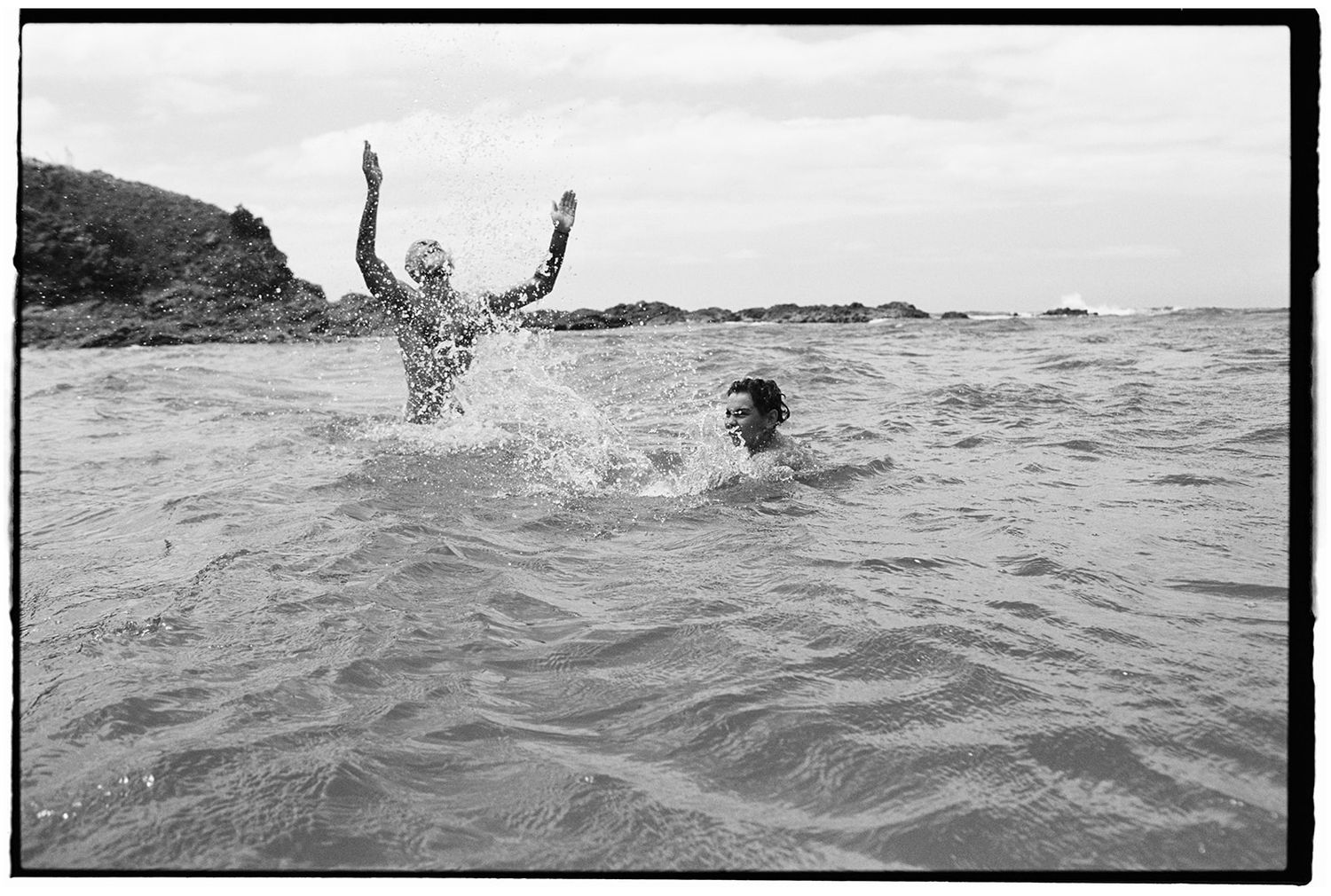 Two people splashing and swimming in ocean waves near rocky coastline in black and white photo.