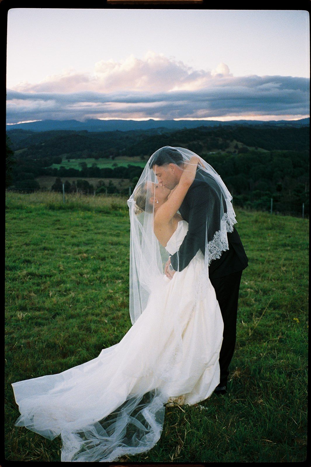Bride and groom sharing a romantic kiss under a lace veil at sunset on a green hillside.