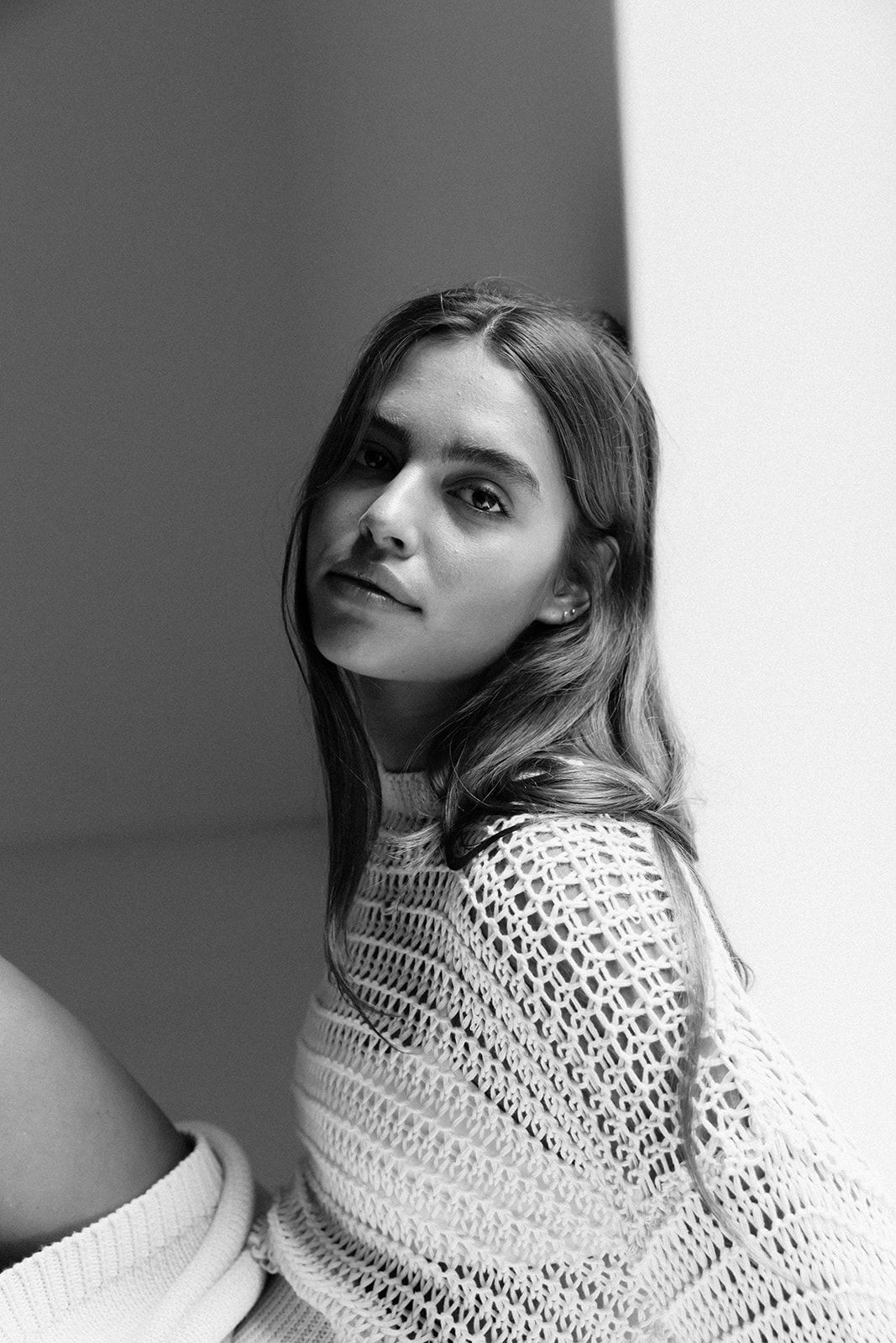 Black and white portrait of a young woman in a crochet sweater, sitting against a wall, looking at the camera.