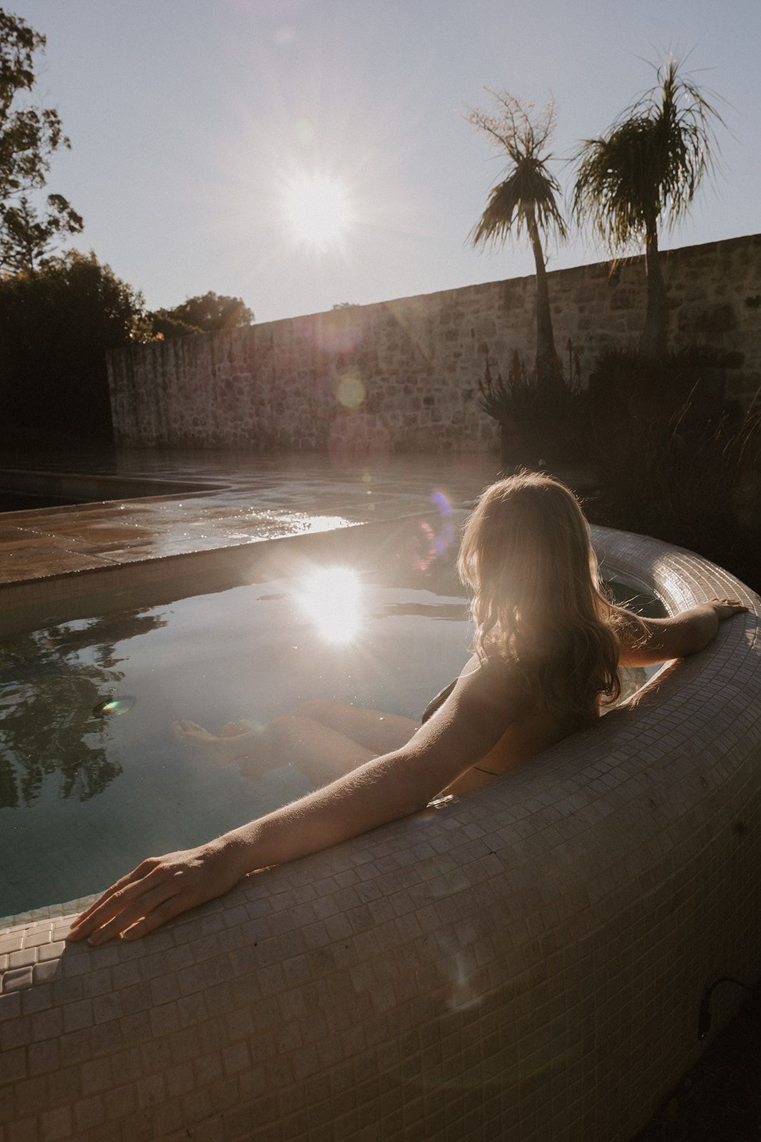 Woman relaxing in a sunlit outdoor pool surrounded by palm trees and stone walls during golden hour.