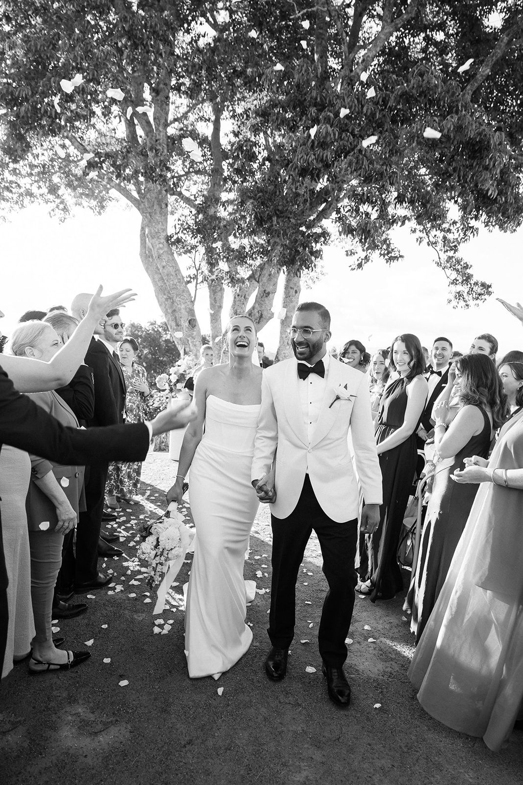 Wedding party in formal attire standing in a line outdoors under shady trees performs synchronized dance moves.