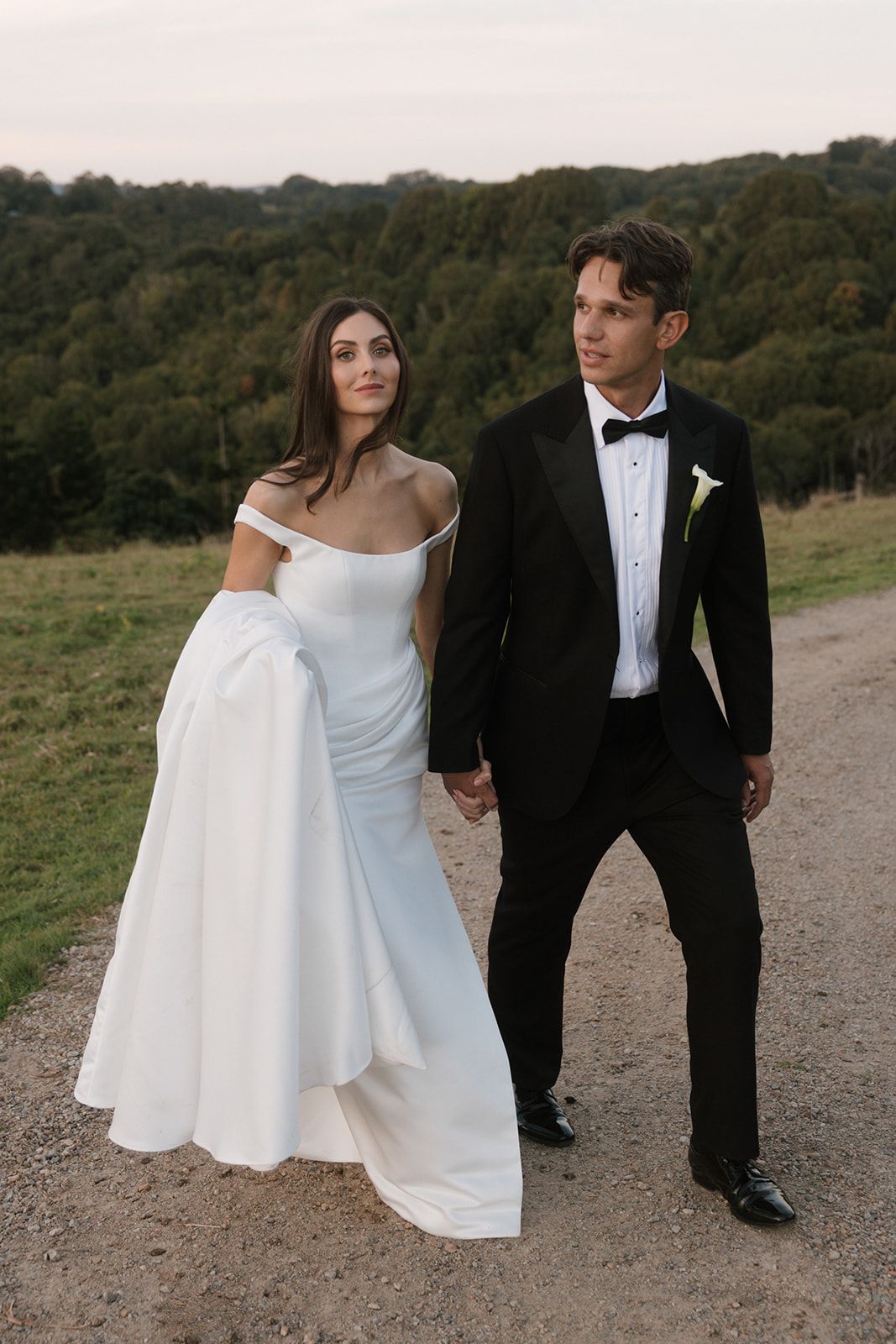 A bride in a white off-shoulder gown holds hands with a groom in a black tuxedo on a scenic countryside path.