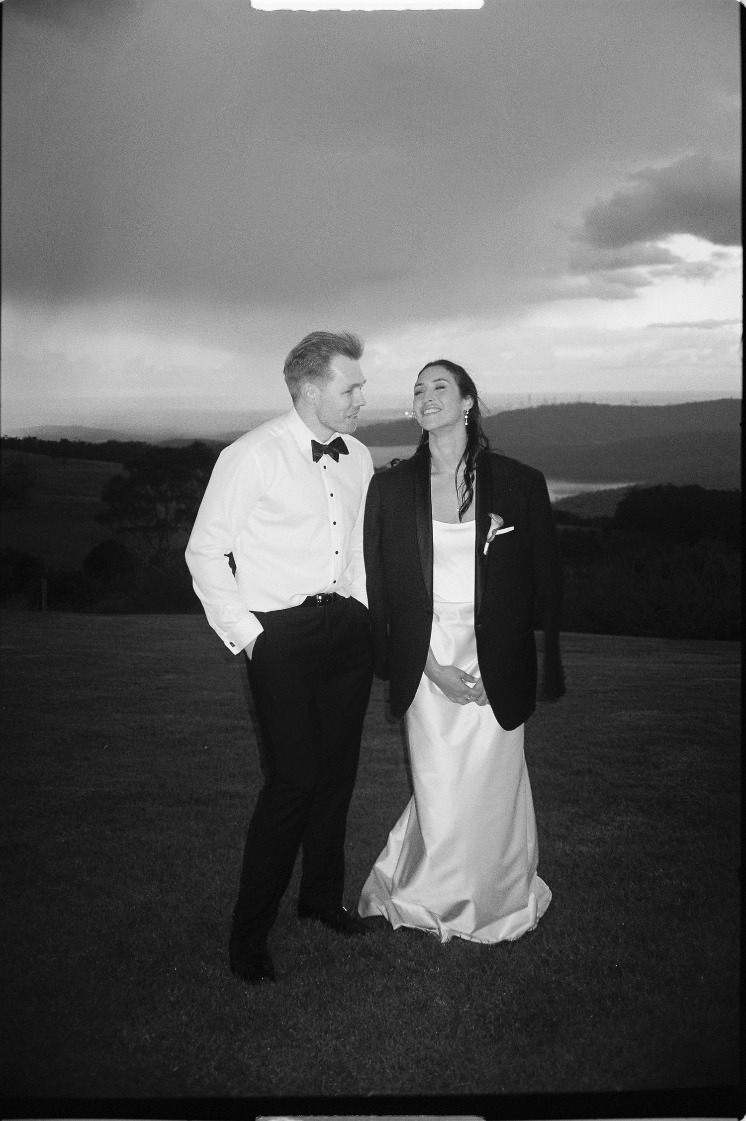 A black and white wedding photo shows two people in formal attire embracing outdoors against a scenic mountain backdrop.