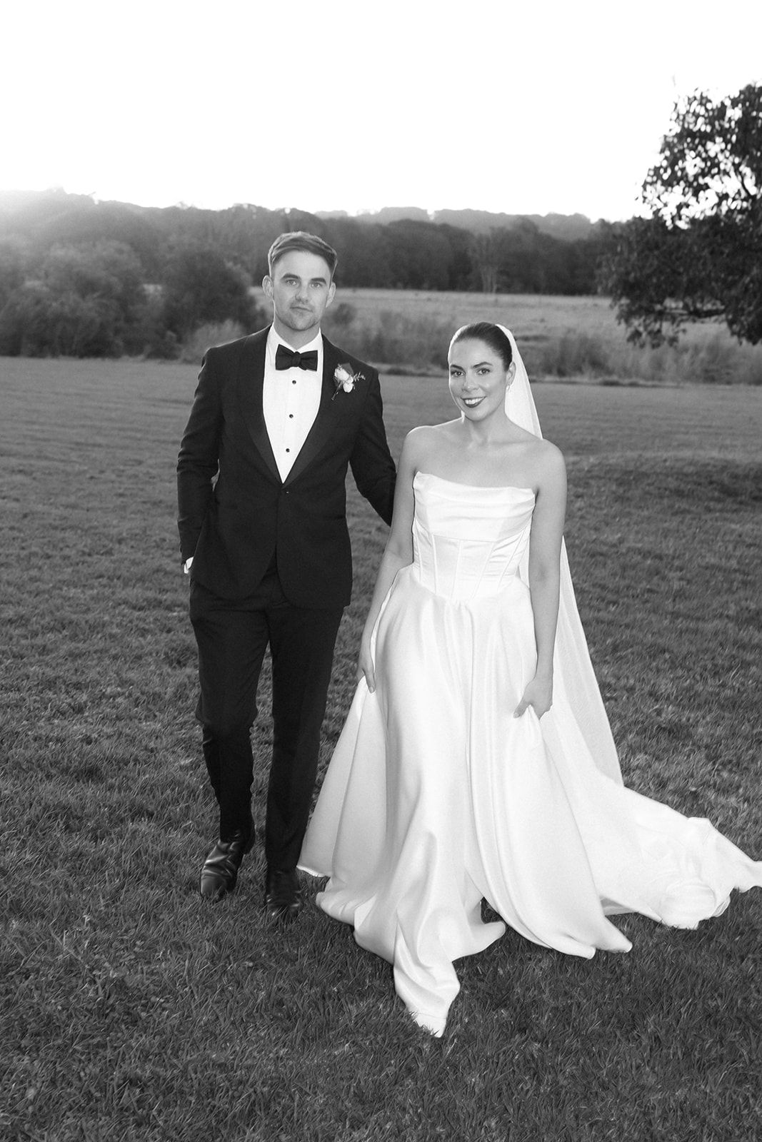 Black and white photo of a bride and groom posing in a field at sunset, wearing elegant wedding attire.