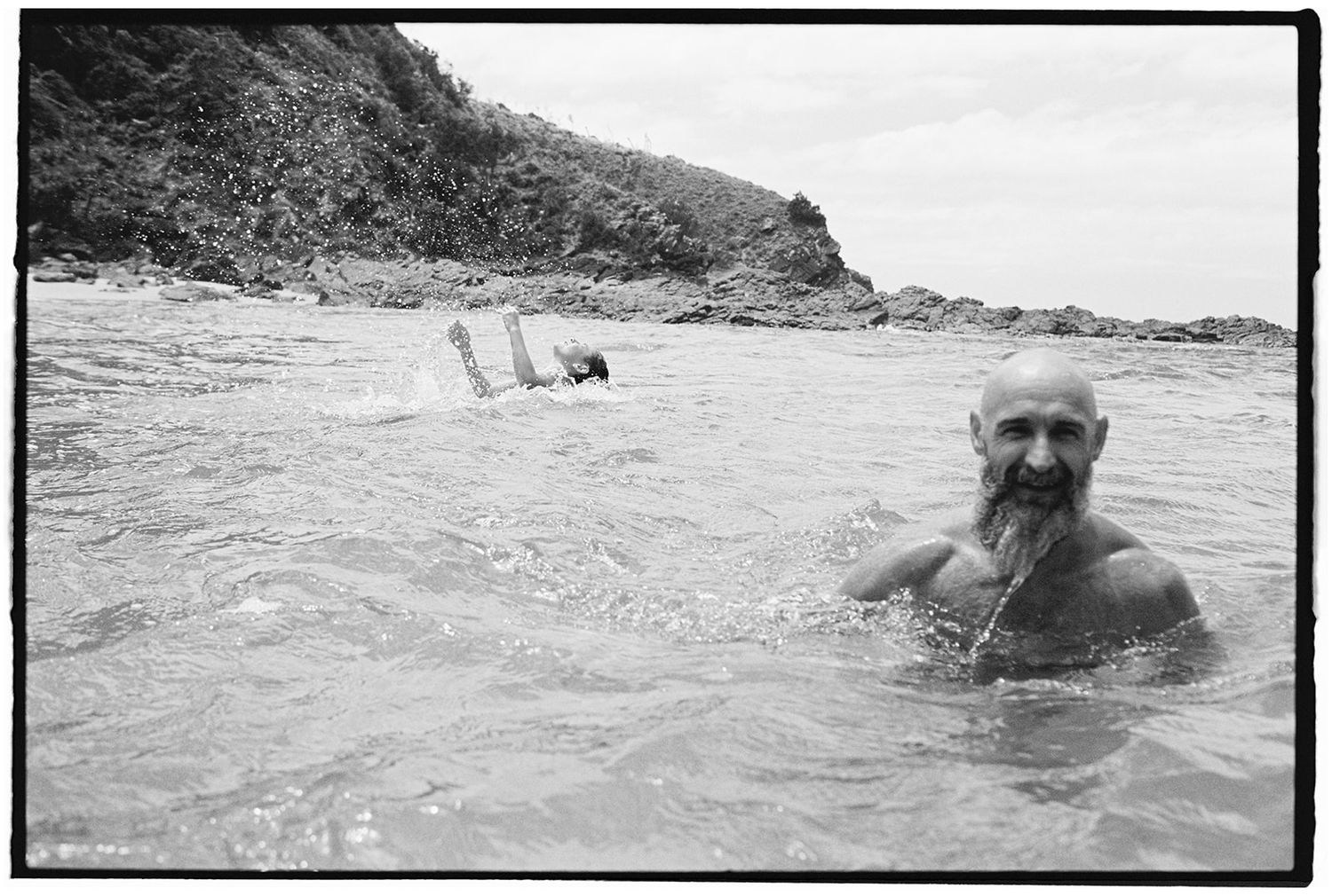 Black and white photo of an older bearded man swimming in ocean near rocky coastline with another swimmer in background.