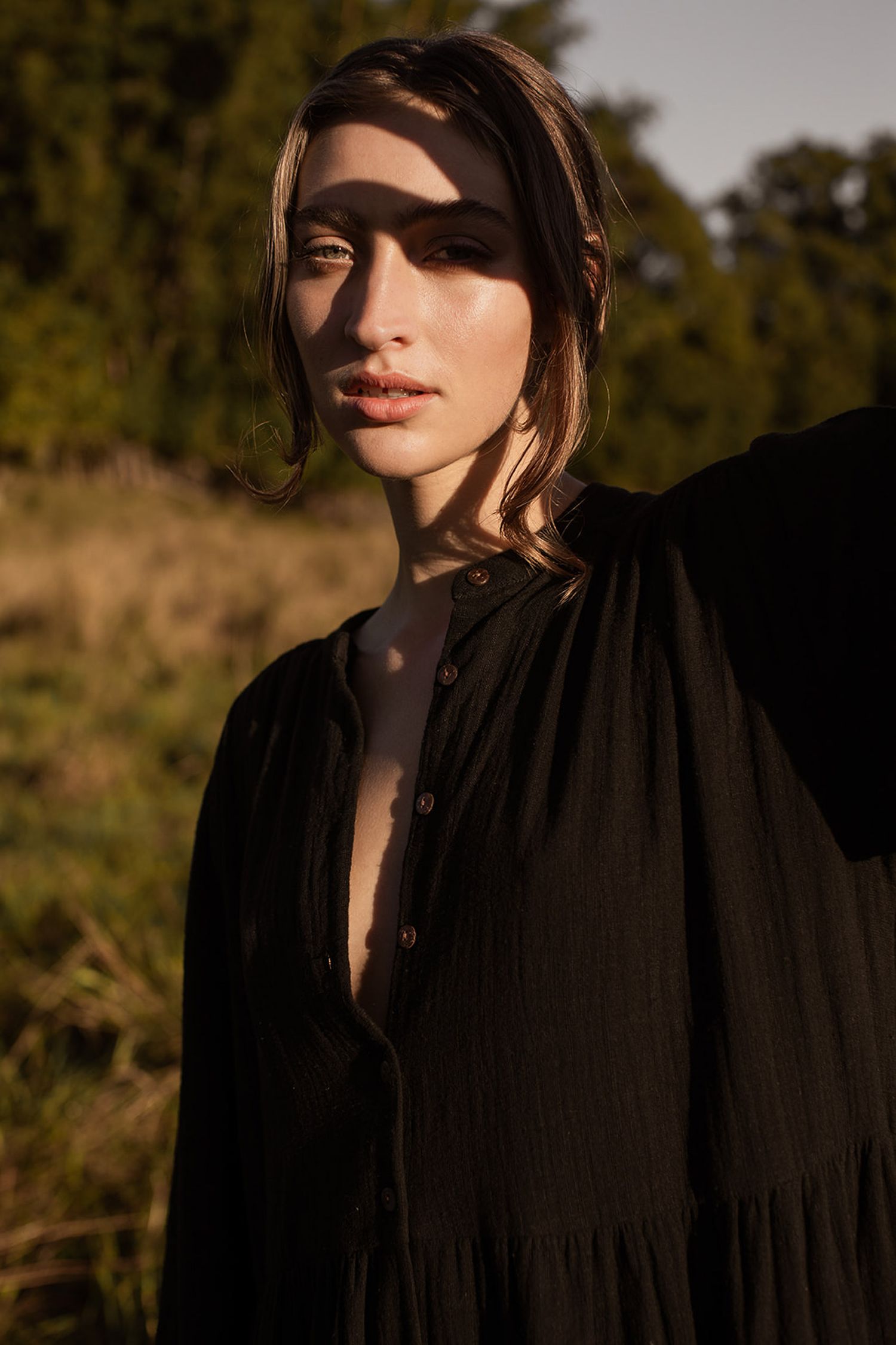 Artistic portrait of someone in a black shirt against a sunlit outdoor background with dramatic shadows and lighting.