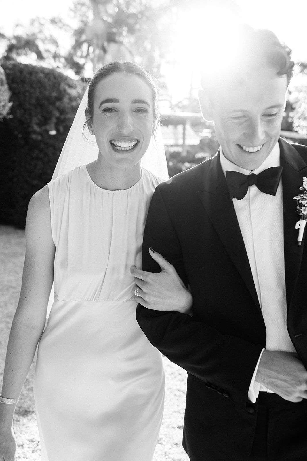 Happy newlyweds laugh together in black and white photo with sunlight behind them, bride in sleek dress and groom in tuxedo.