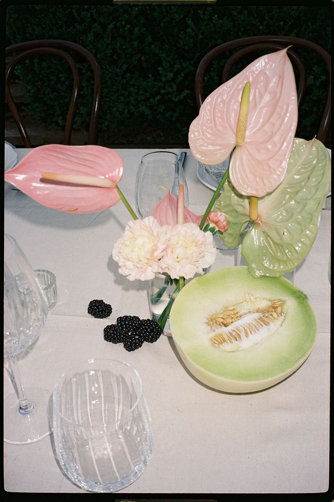 Elegant table setting with pink anthuriums, white carnations, honeydew melon, blackberries, and crystal glassware on white linen.