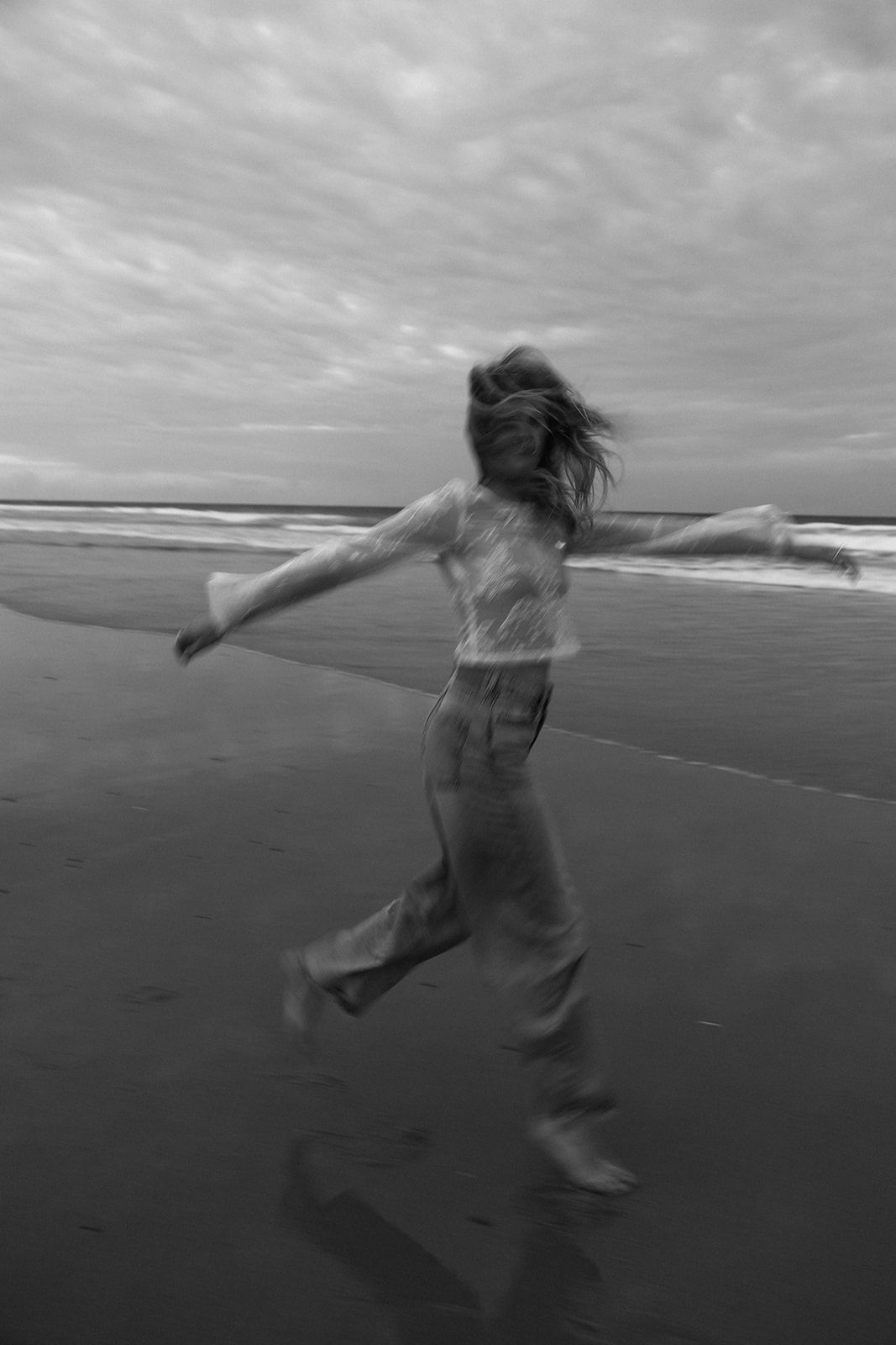 A silhouette dances freely on a cloudy beach, hair flowing in the wind in black and white photography.