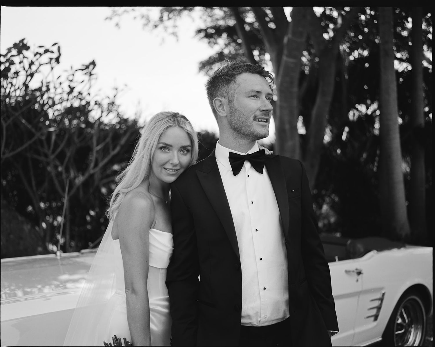 Black and white photo of a bride and groom in wedding attire smiling together outdoors near a vintage car.