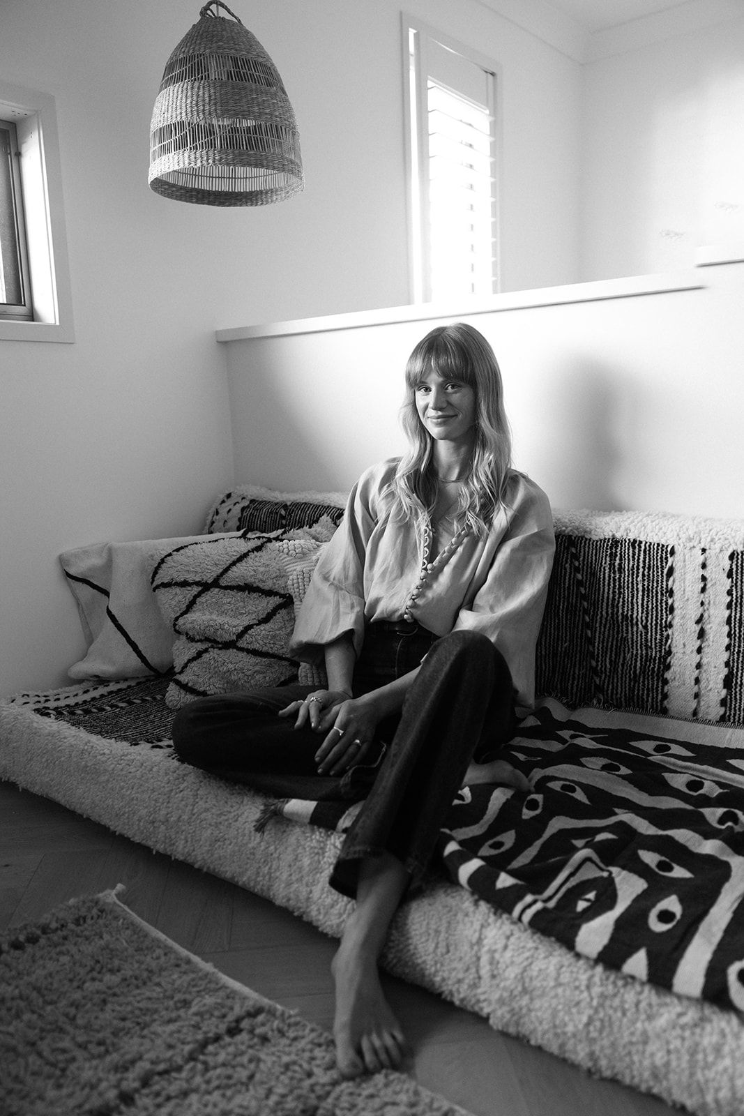 Woman sitting barefoot on a boho-style daybed with patterned cushions and throws in a bright minimalist room.