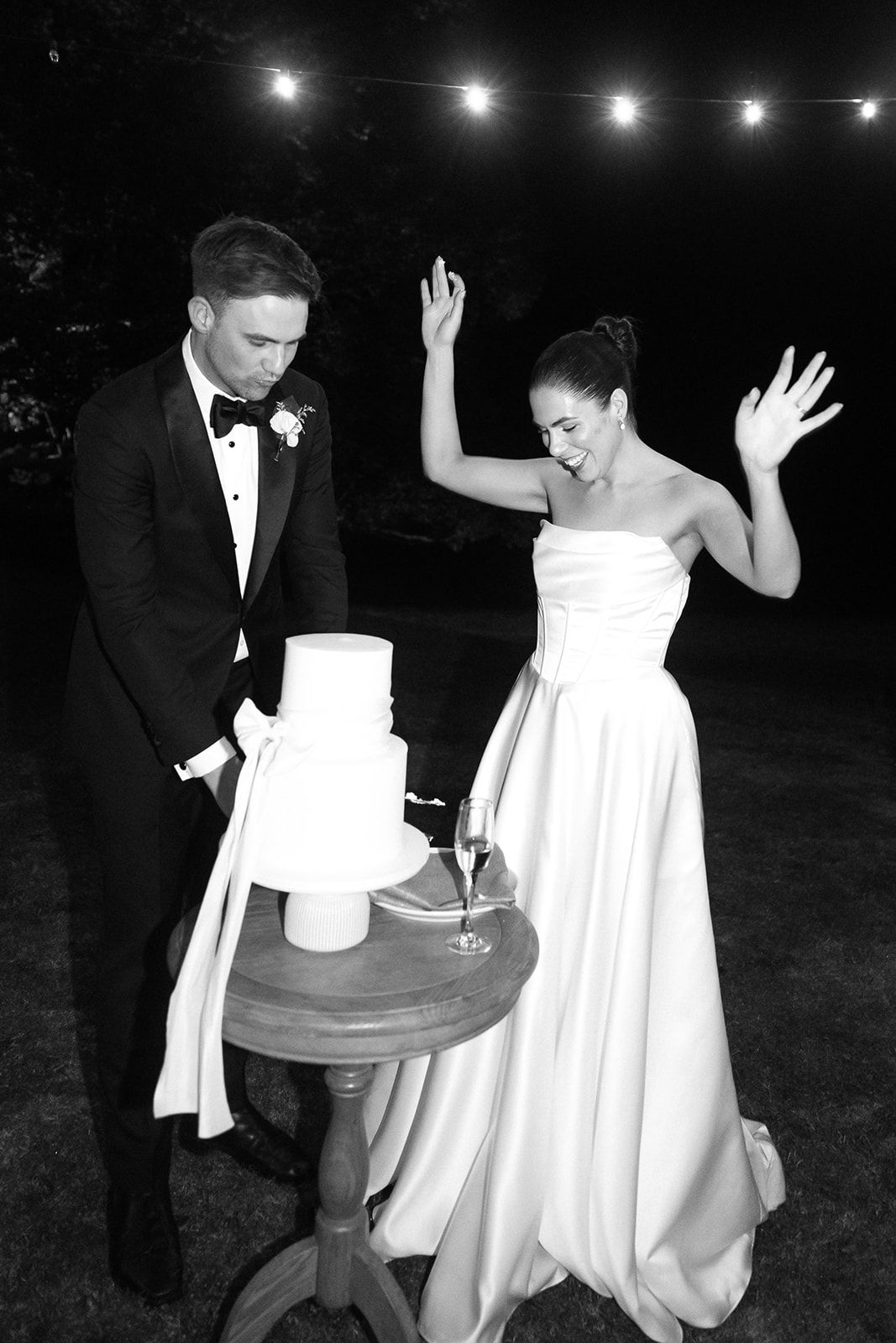 Black and white photo of newlyweds celebrating at their wedding reception with string lights overhead.