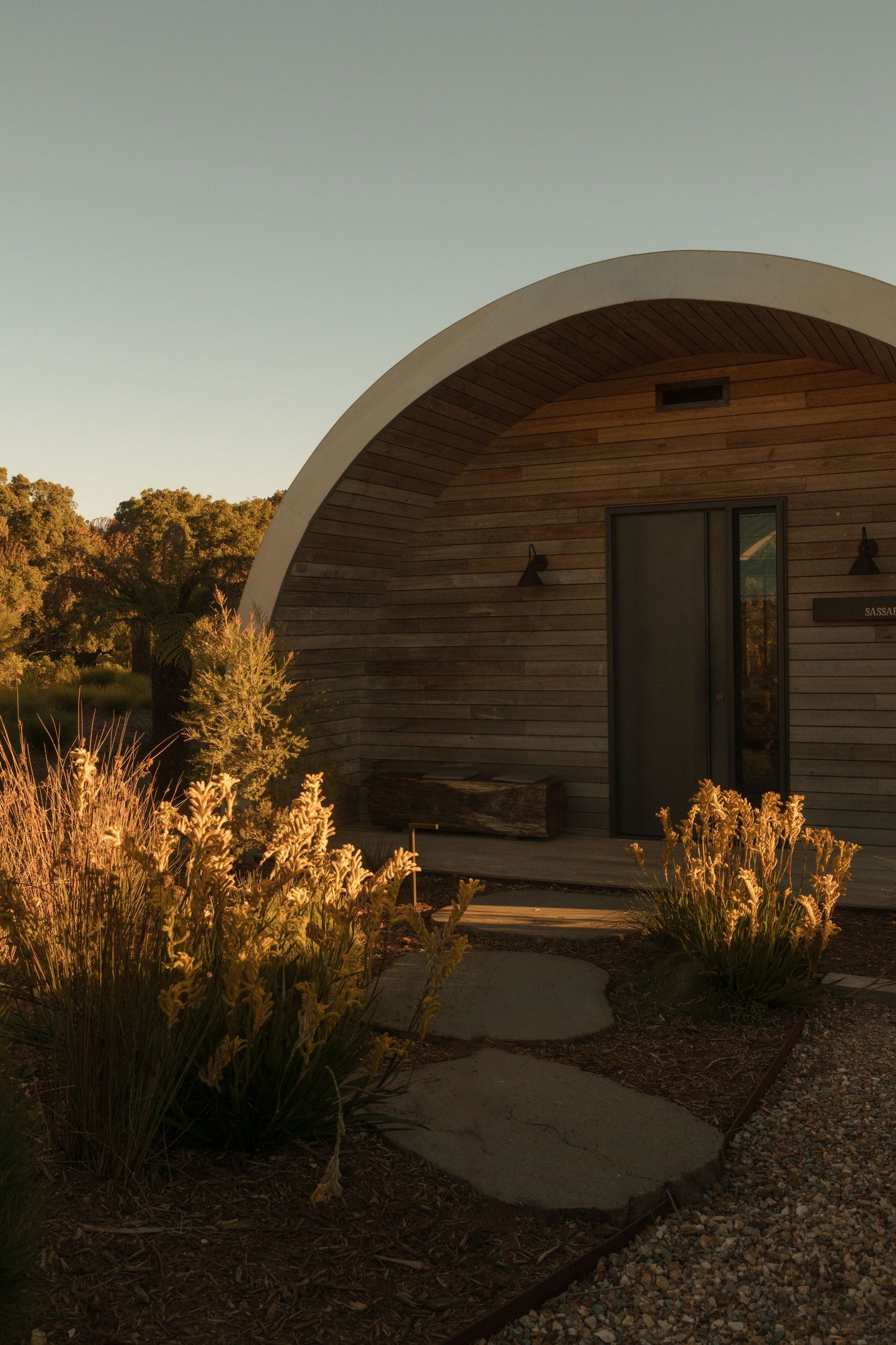 Curved wooden cabin with arched roof, stone path, and golden wild grasses at sunset in a natural landscape.