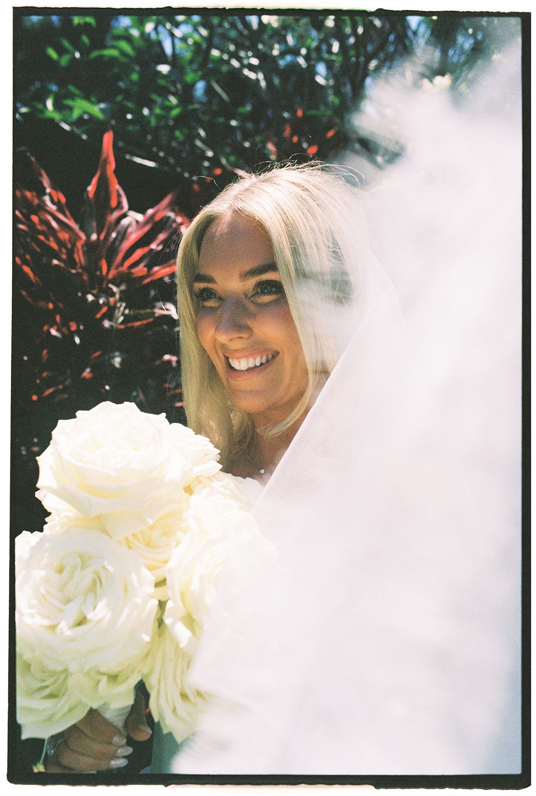Smiling blonde bride holding white rose bouquet, partially veiled, with lush tropical greenery background.