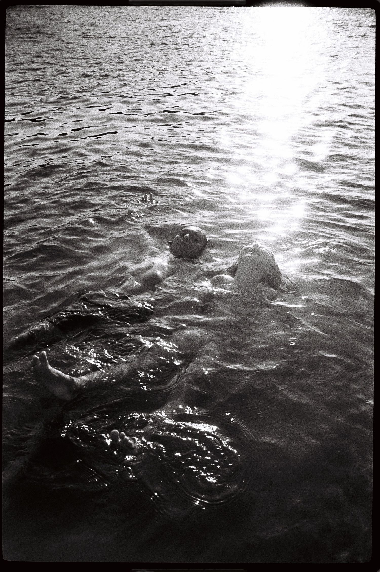 Black and white photo of a person swimming in sunlit rippling water, with bright light reflecting on the surface.