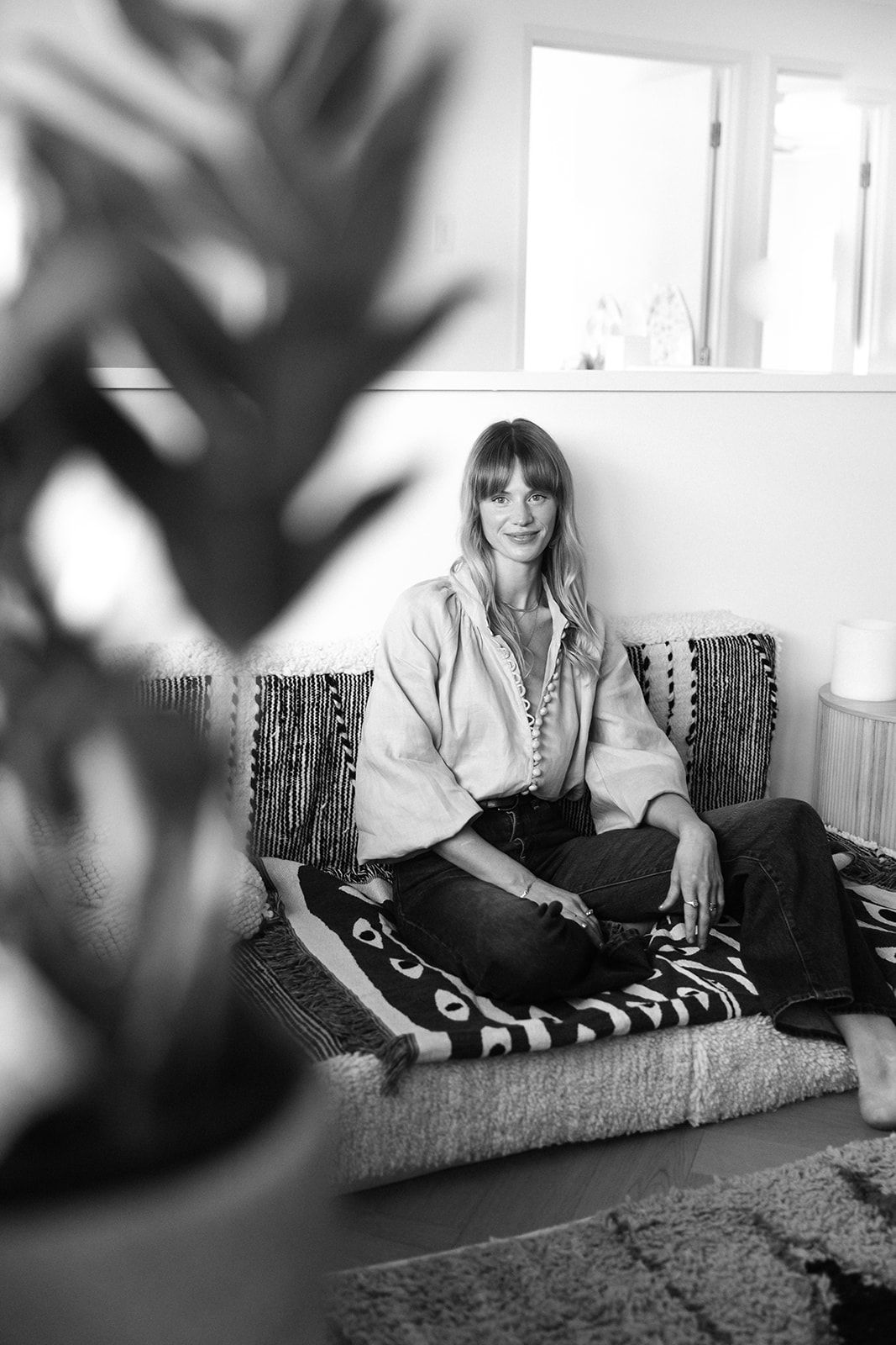 Black and white photo of a woman sitting cross-legged on a patterned sofa, smiling, with a plant in the foreground.