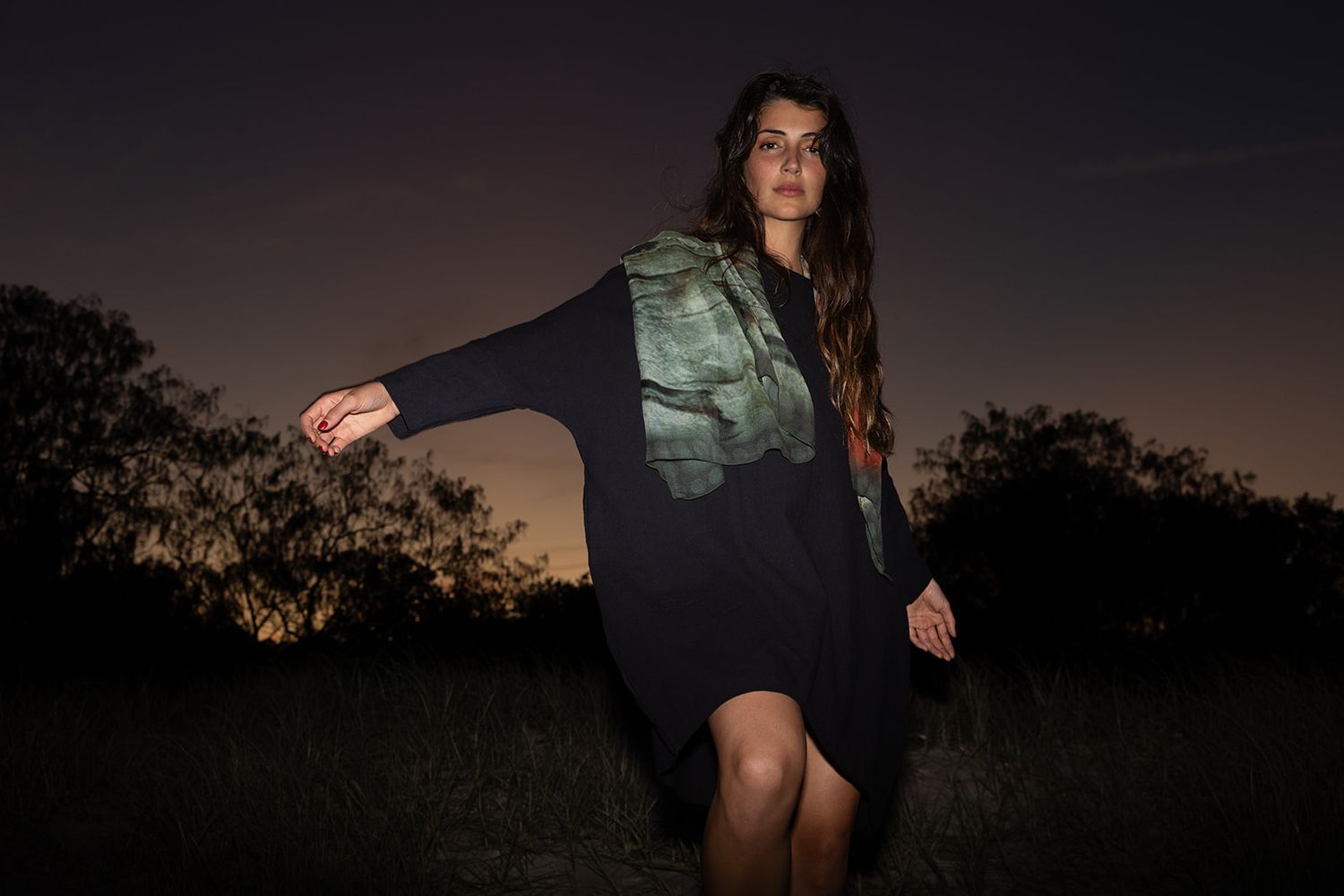 Woman in black dress and olive vest poses dramatically outdoors at dusk with dark sky and trees behind her.