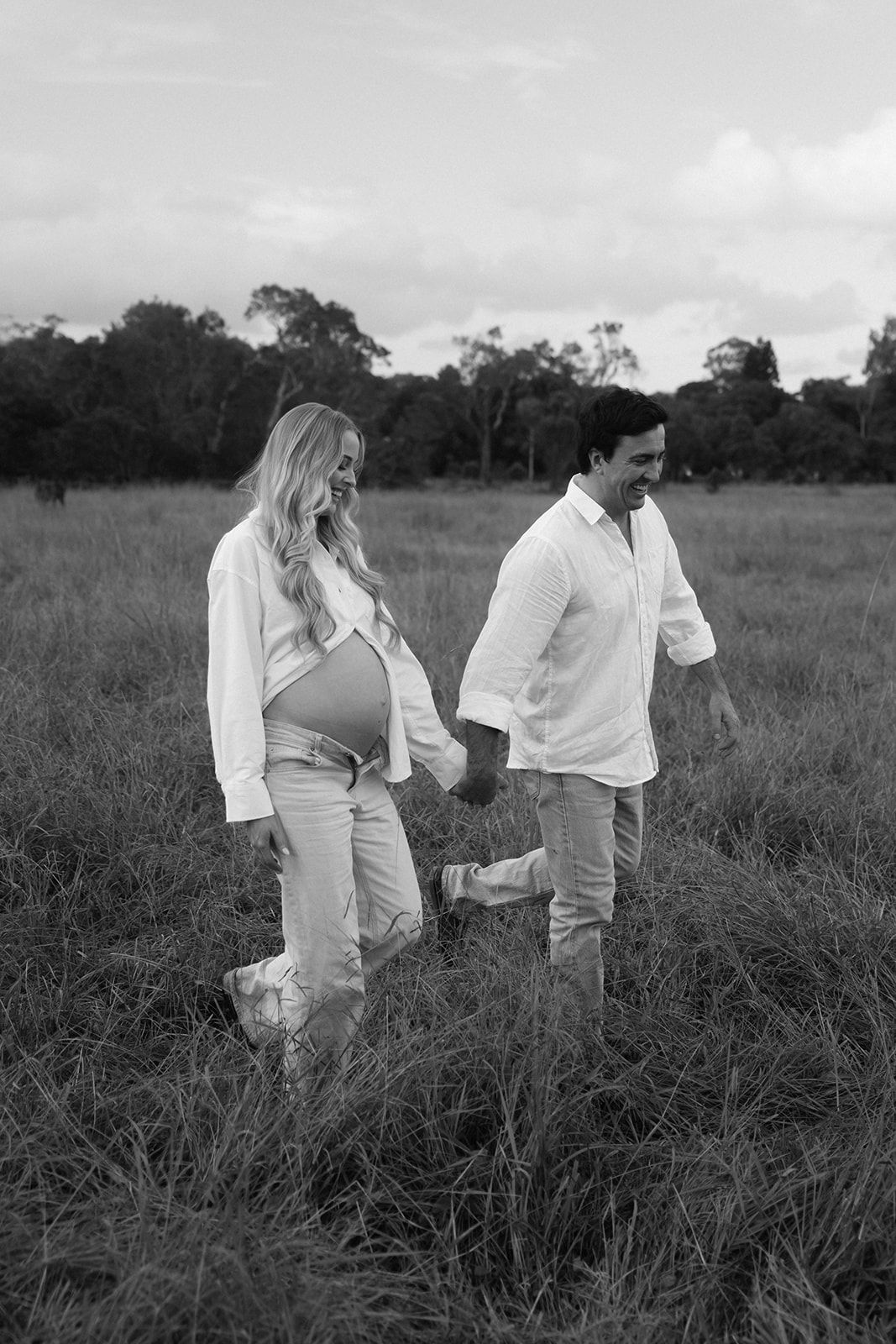 Black and white photo of a pregnant couple holding hands while walking through a grassy field outdoors.
