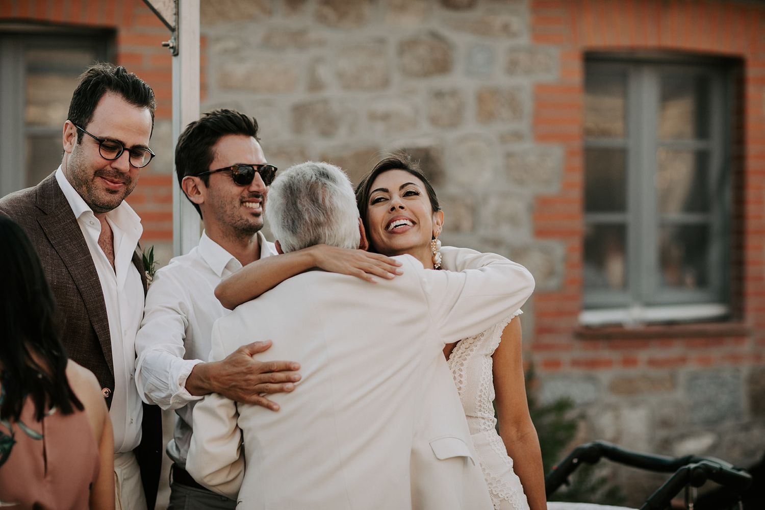 Wedding guests share laughter and emotional moments outside a rustic brick building during a sunny celebration.
