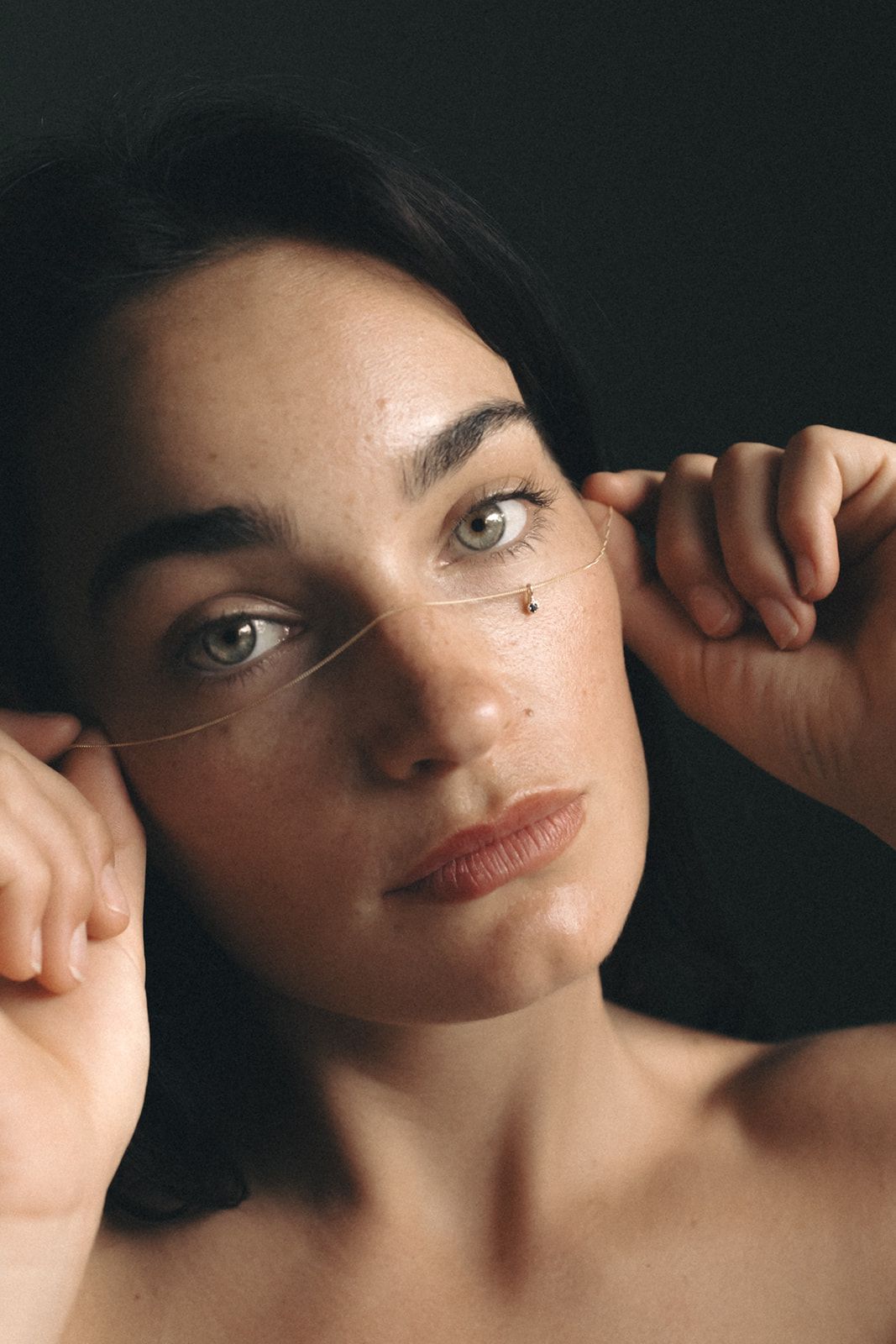Close up portrait with dramatic lighting against dark background showing bare shoulders and hands framing eyes.