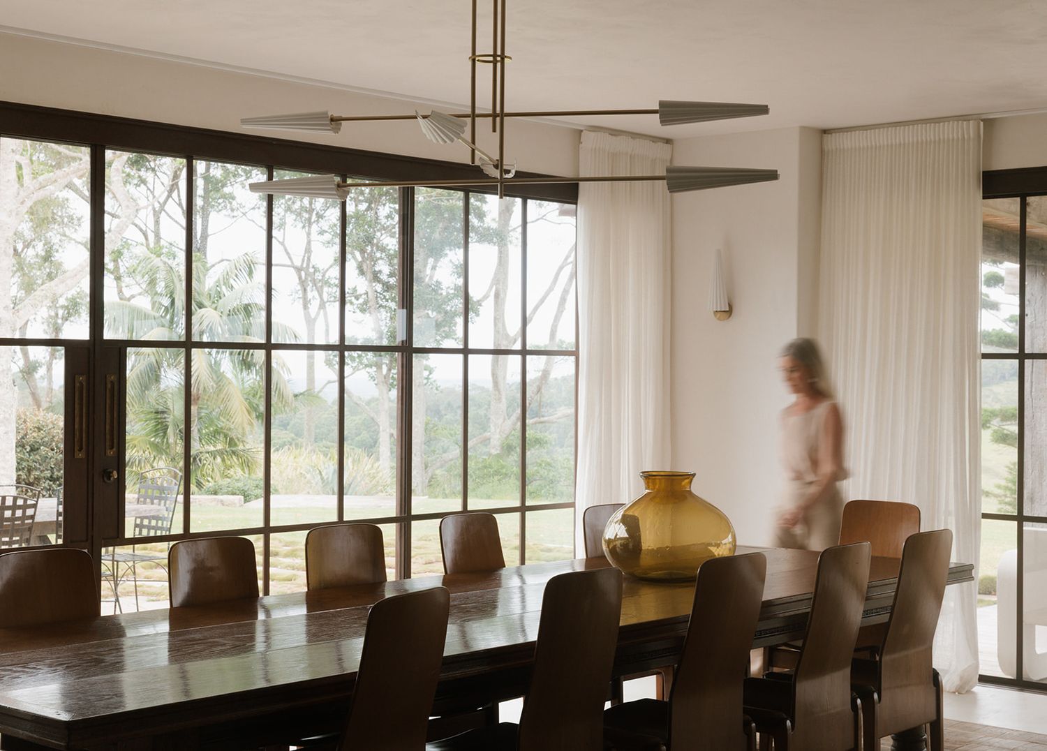 Elegant dining room with large windows, long wooden table, leather chairs, yellow vase centerpiece, and sheer white curtains.