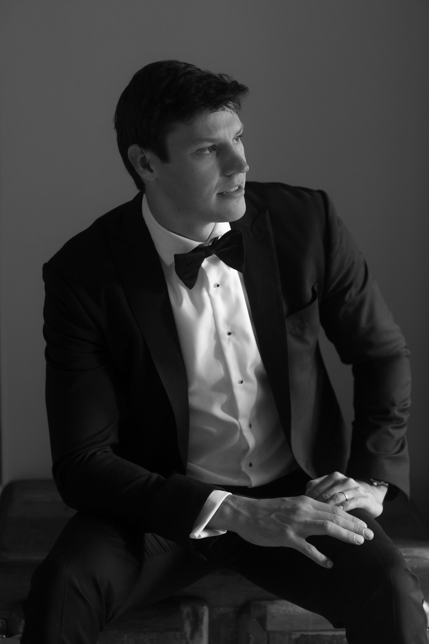 Black and white portrait of a young man in a tuxedo with bow tie, sitting and gazing thoughtfully to the side.