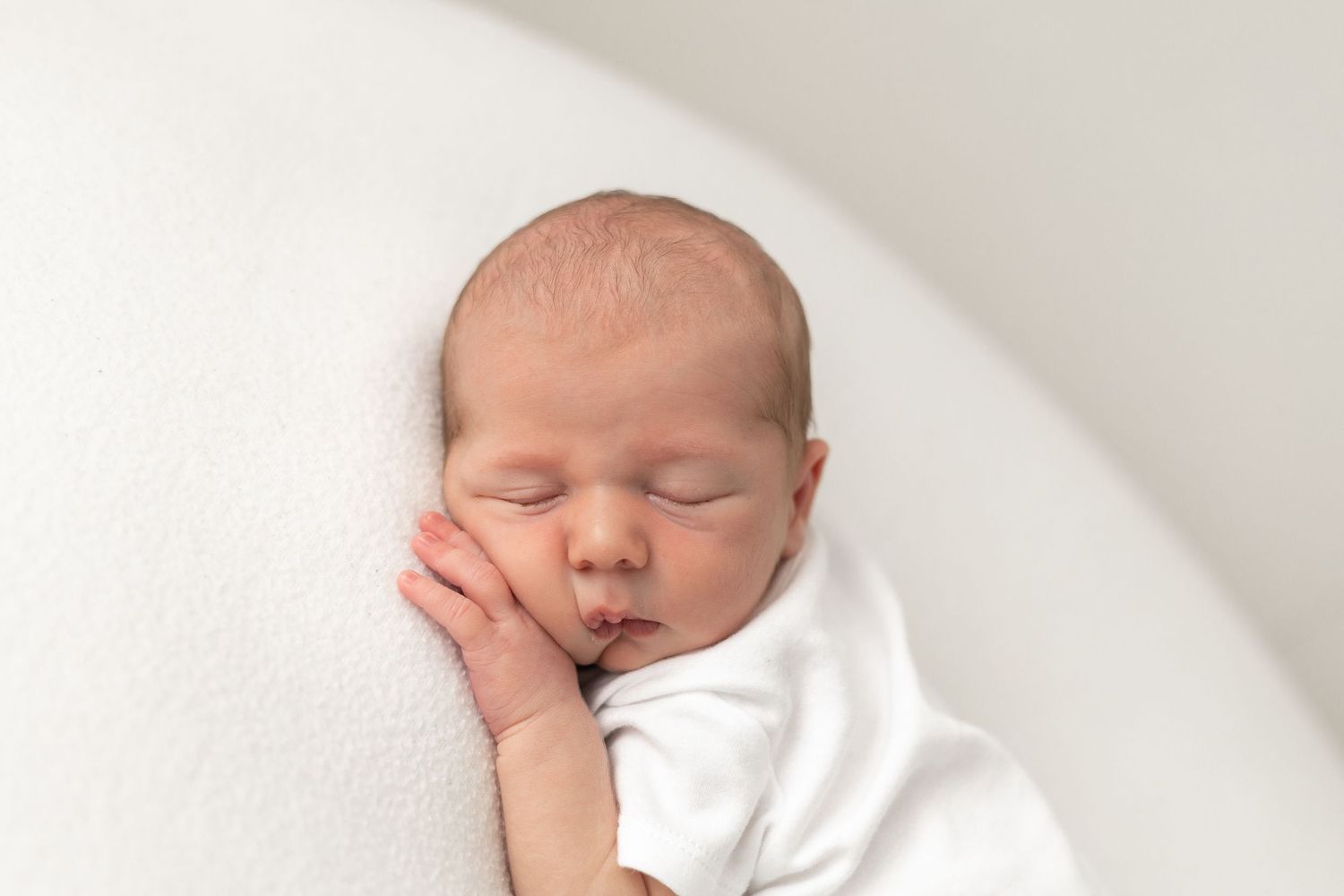 Peaceful newborn baby sleeping on white blanket, wrapped in white cloth with tiny hands near face.