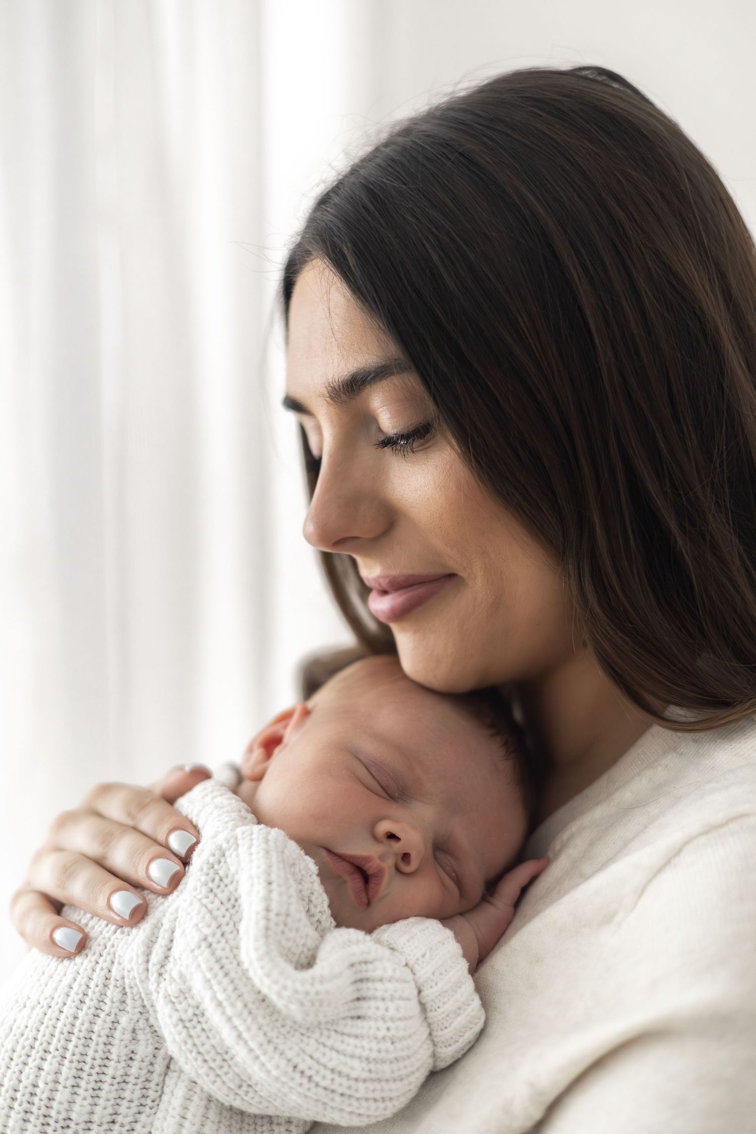 A young mother with dark hair smiles tenderly while cradling her sleeping newborn baby dressed in white.