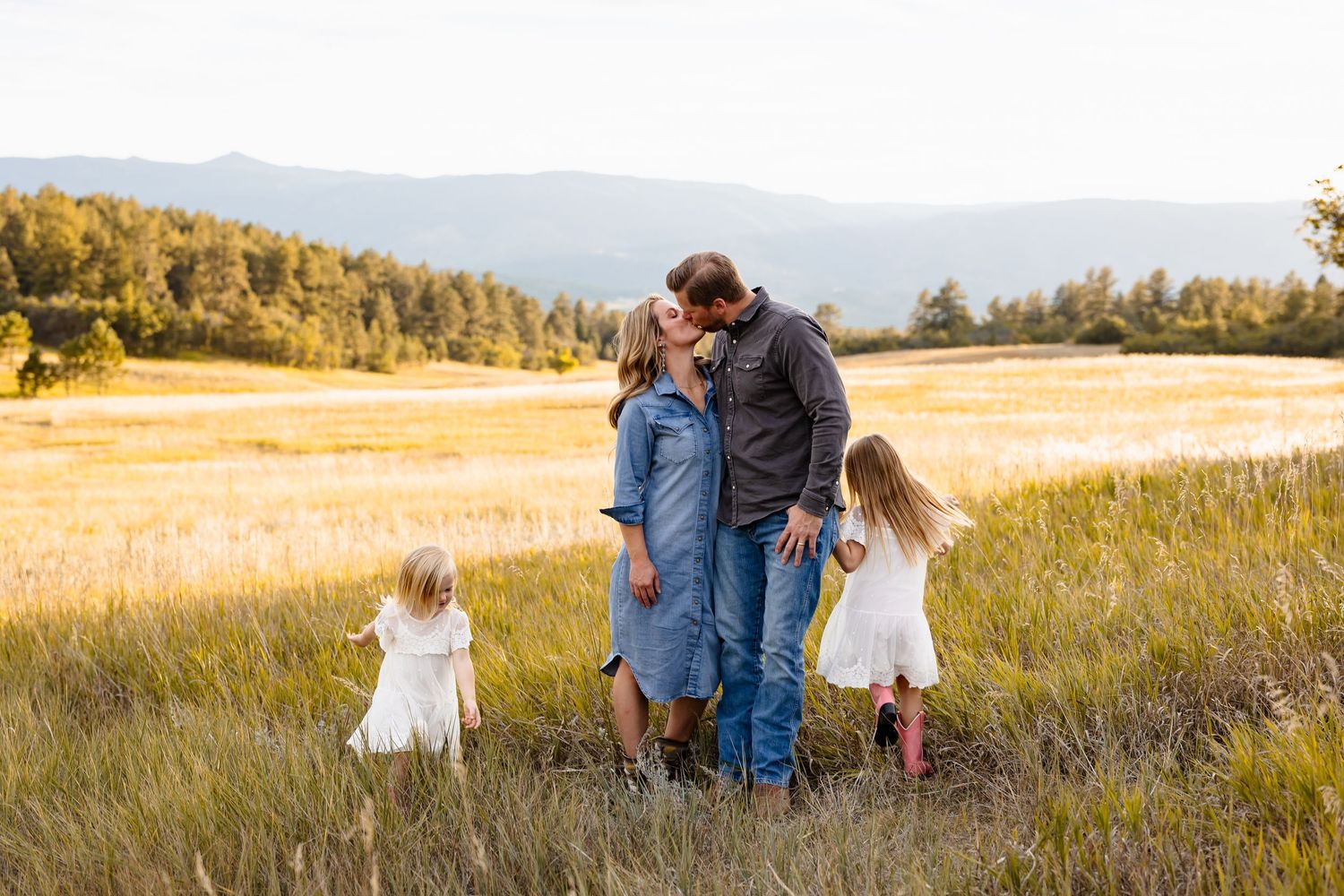 Couple sharing a romantic kiss in a golden field at sunset with two young daughters nearby.