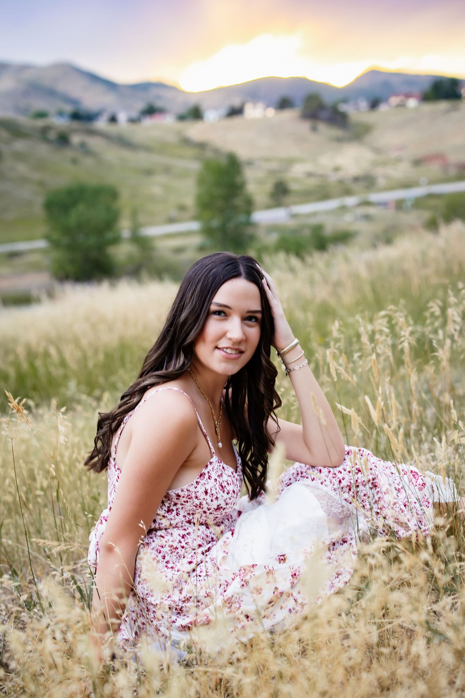 Young woman in floral sundress sitting in golden meadow grass at a colorful mountain sunset.