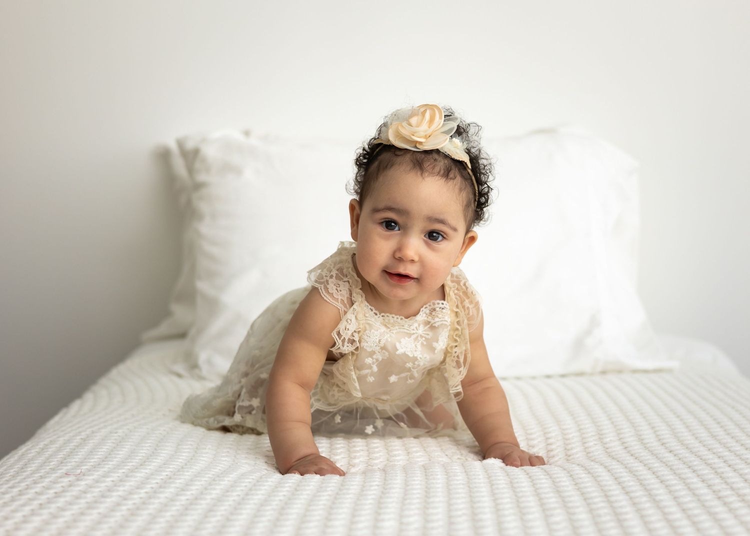 Adorable baby girl in cream lace dress and floral headband crawling on white bed during professional photo session.