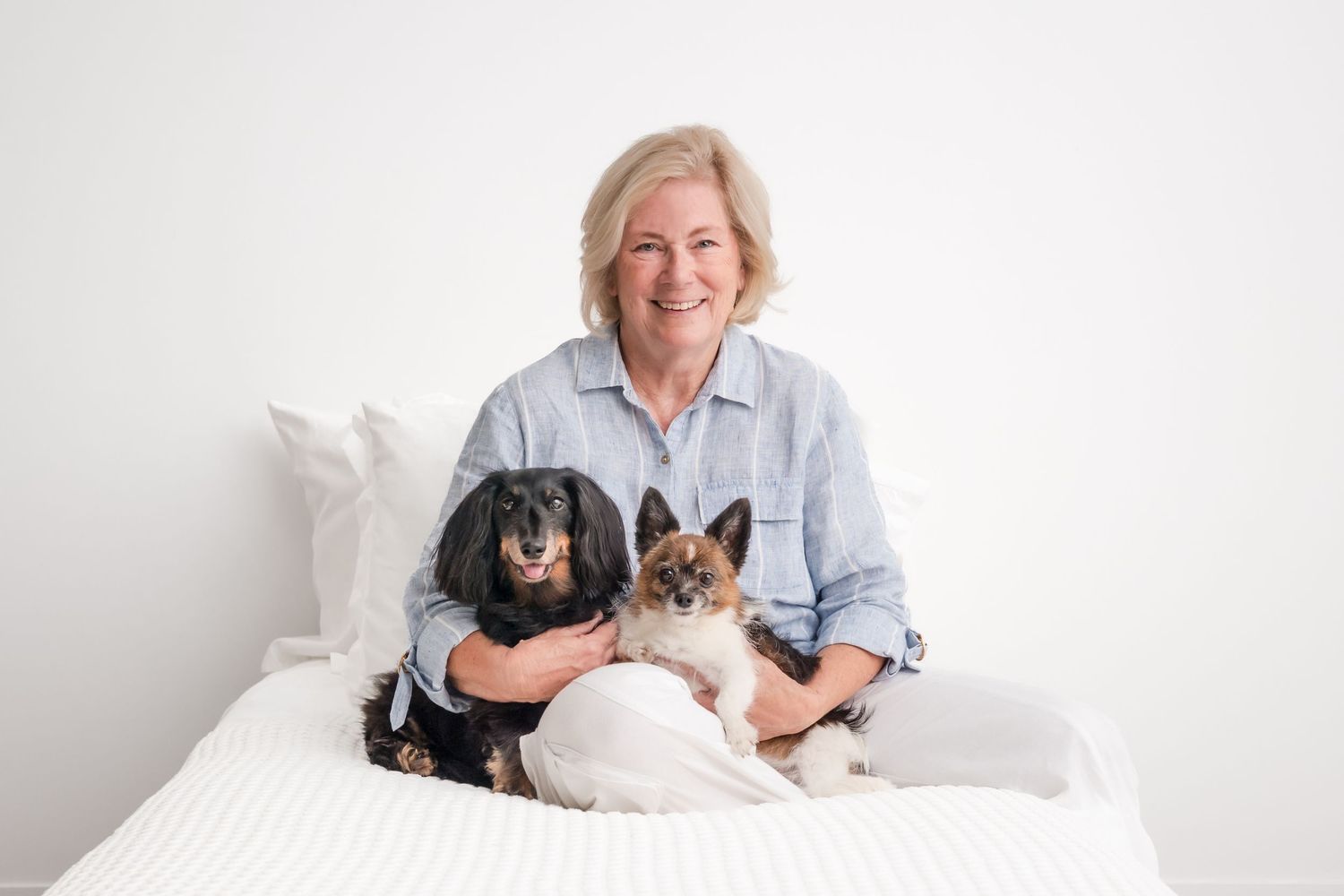 Smiling woman sitting on white bed with black Cocker Spaniel and Siamese cat, bright minimalist background.